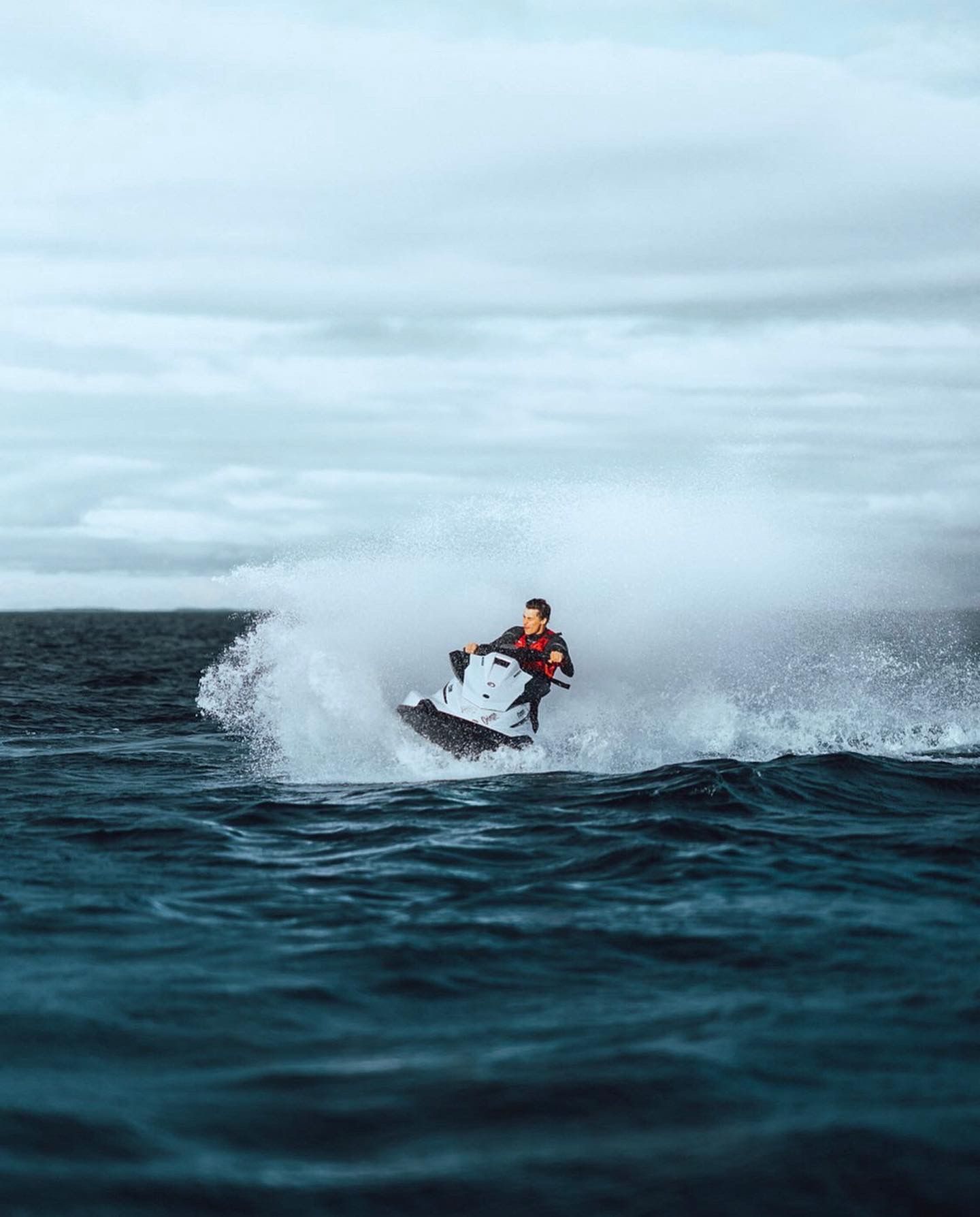 A jet ski rider kicks up spray while jumping a wave on dark blue ocean waters under an overcast sky.