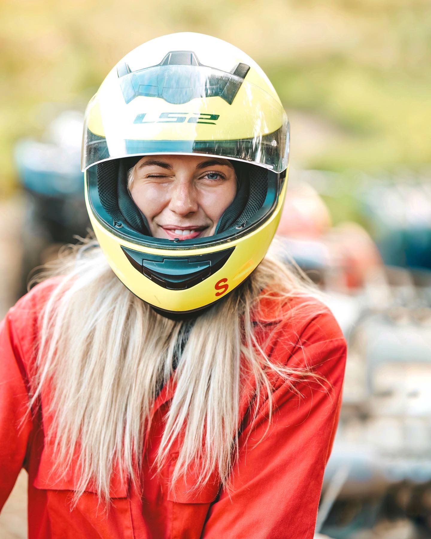 Race car driver wearing bright yellow helmet and red racing suit with long blonde hair smiling.