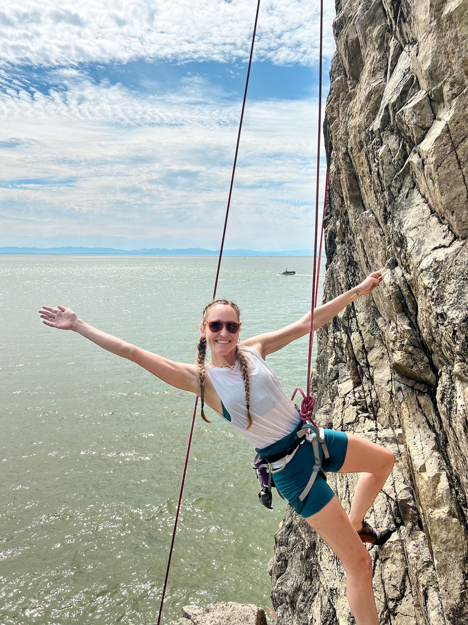 Rock climber in sunglasses and climbing harness poses joyfully while rappelling down a cliff face over sparkling waters.