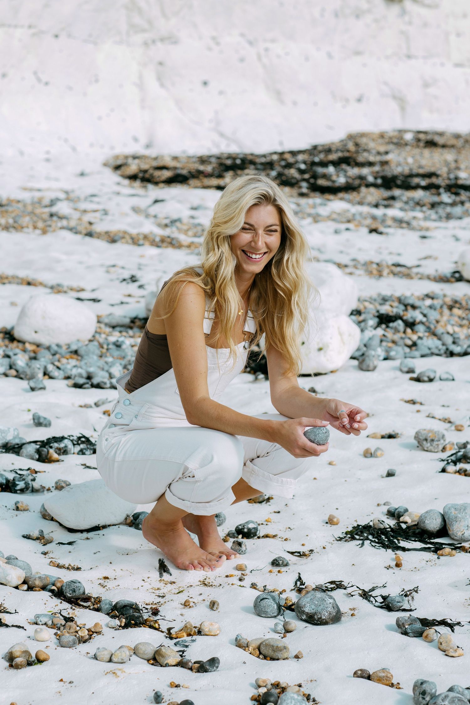 Woman in white dress kneels on the sand at a coastal beach with rocks and stones scattered around.