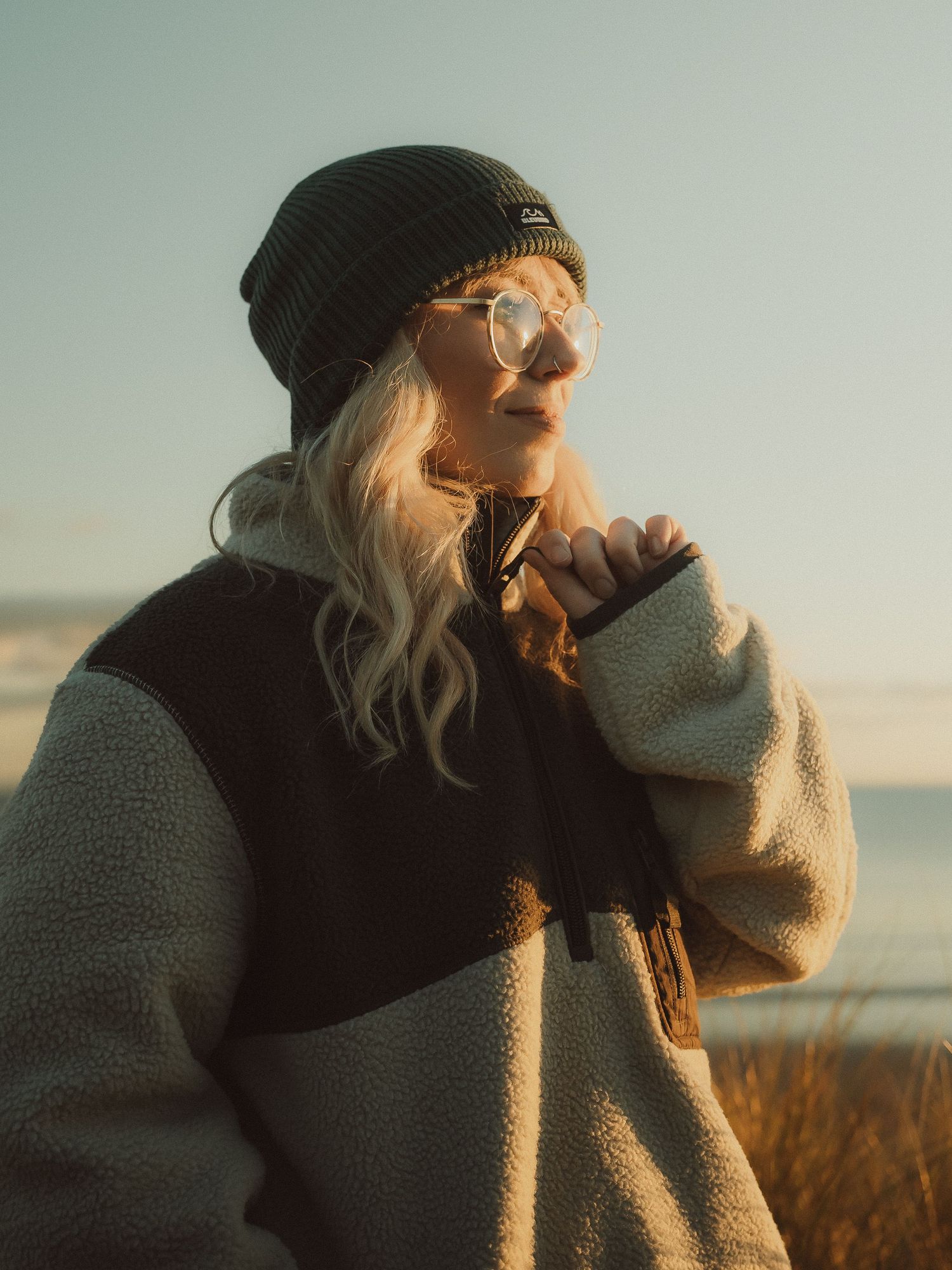 Person in a beanie and cozy sweater laughs while enjoying golden hour sunlight outdoors.