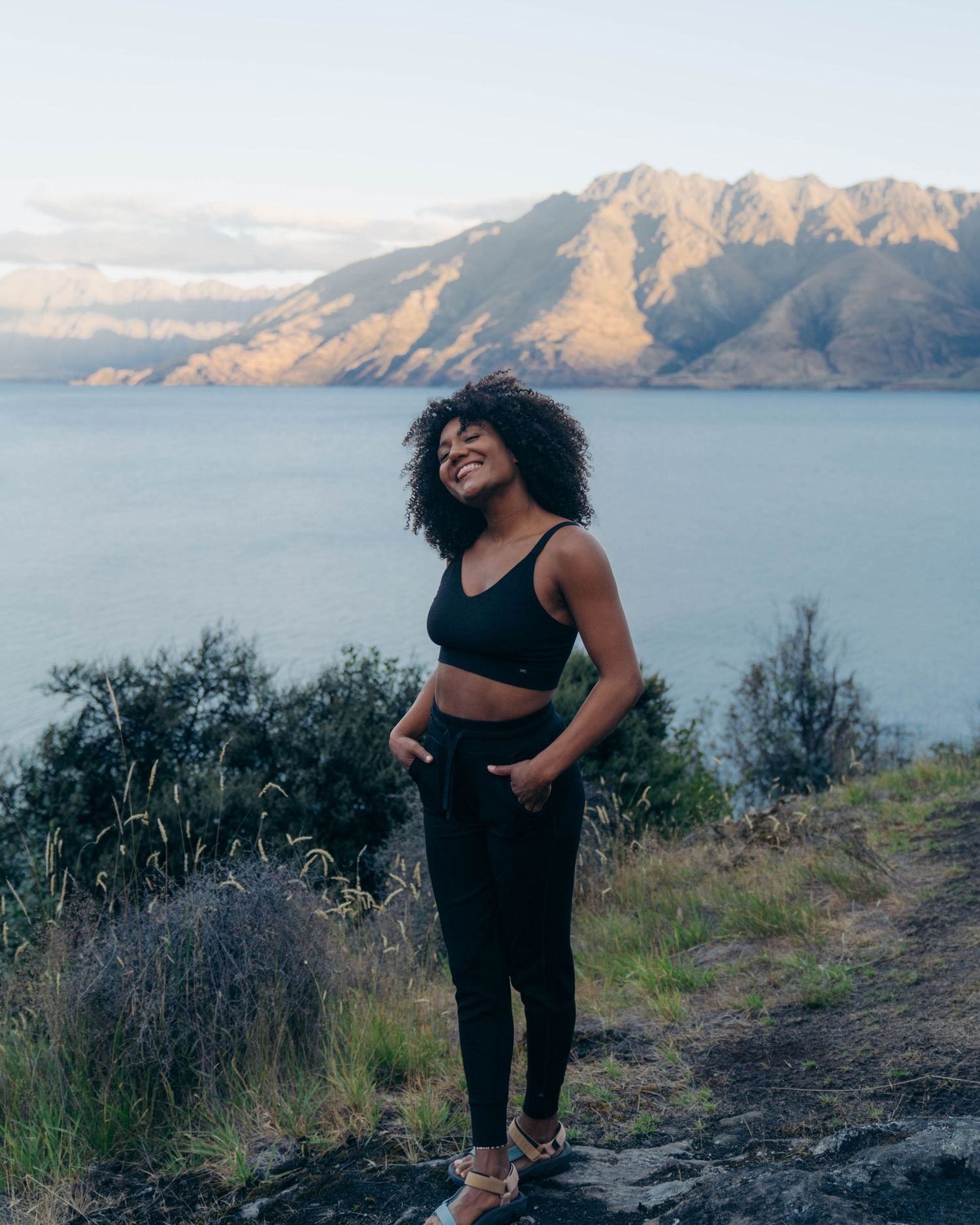 A happy individual in athletic wear poses along a hiking trail overlooking a mountain lake on a sunny day.