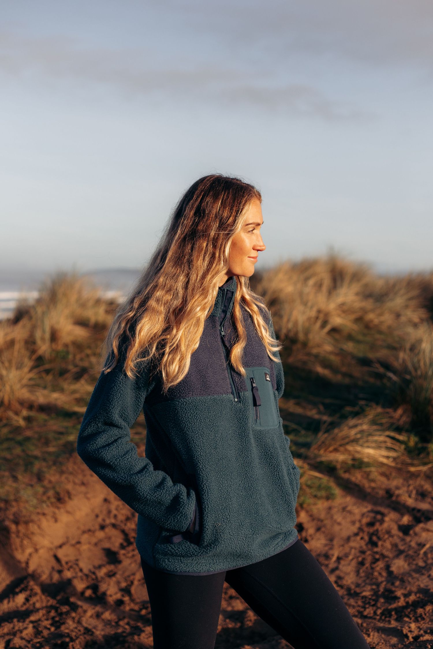 Person in a dark fleece jacket stands against a golden sunset on a desert hillside.