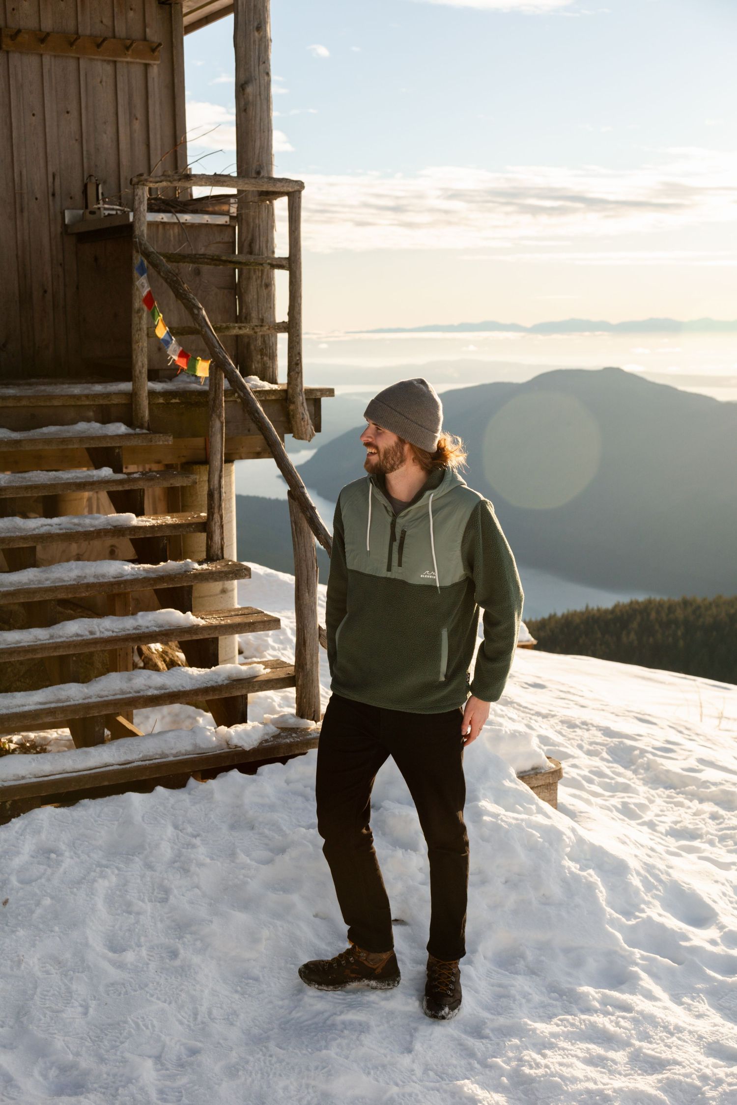 Person in winter attire standing in snow outside a mountain cabin with scenic mountain views at sunset.