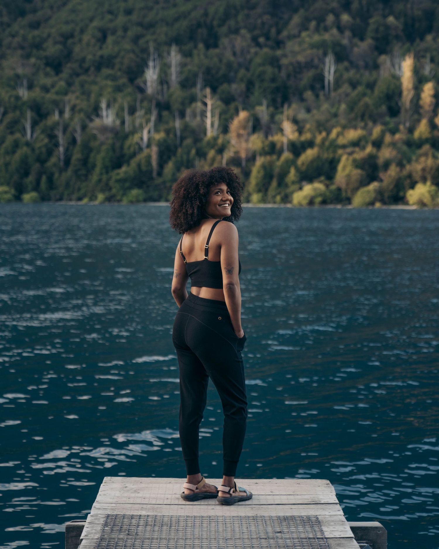 Athletic woman standing on wooden dock by blue mountain lake wearing black athletic wear and sneakers.