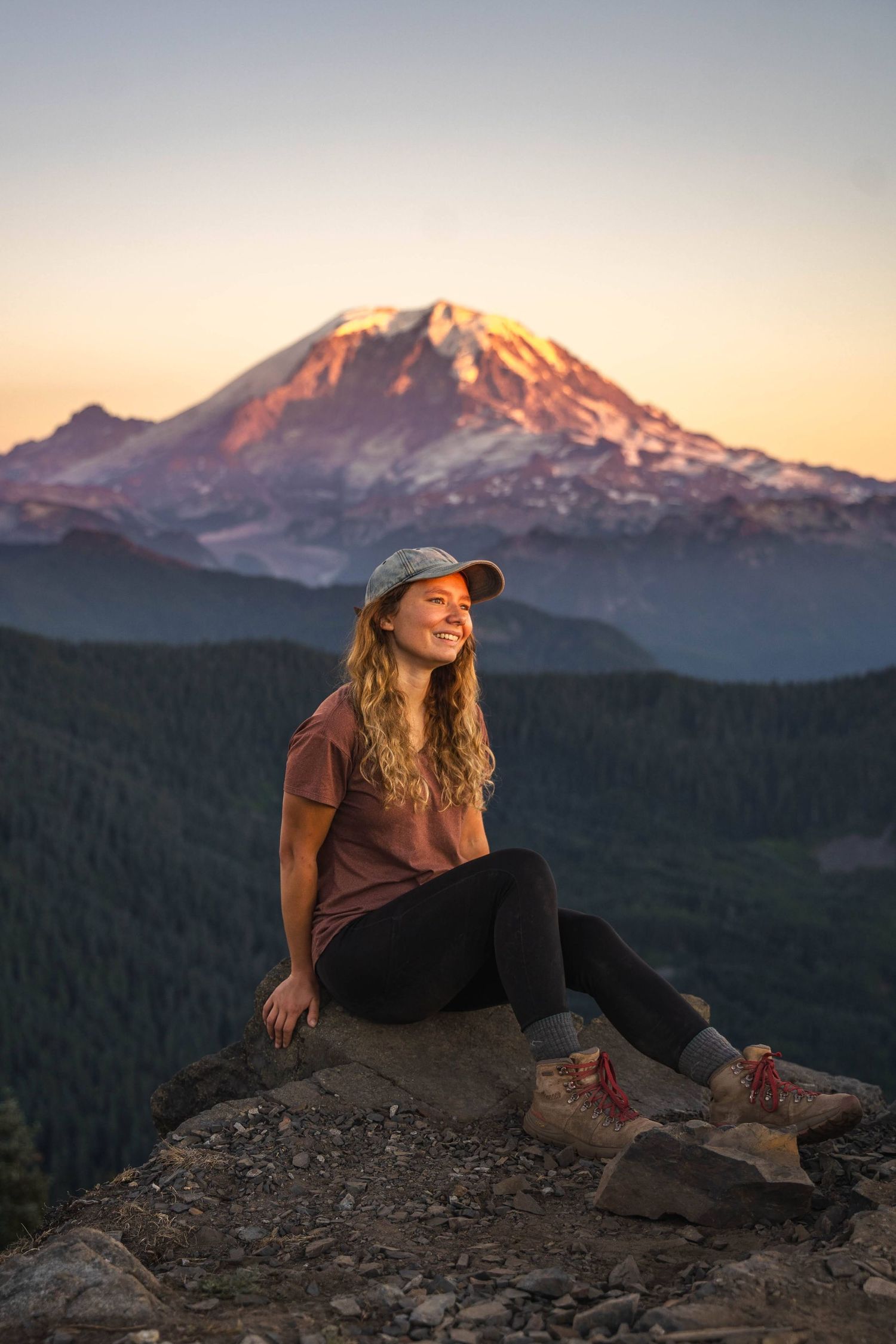 Hiker sits on rocky overlook at sunset with Mount Rainier's snow-capped peak glowing in warm evening light.