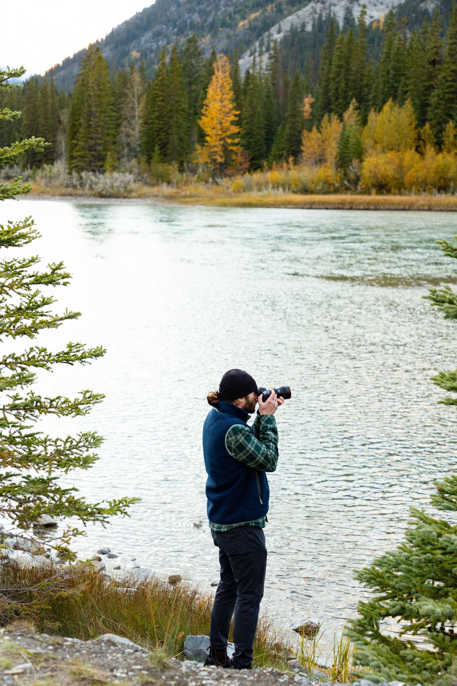A photographer takes pictures by a scenic mountain lake with autumn foliage and evergreen trees along the shoreline.