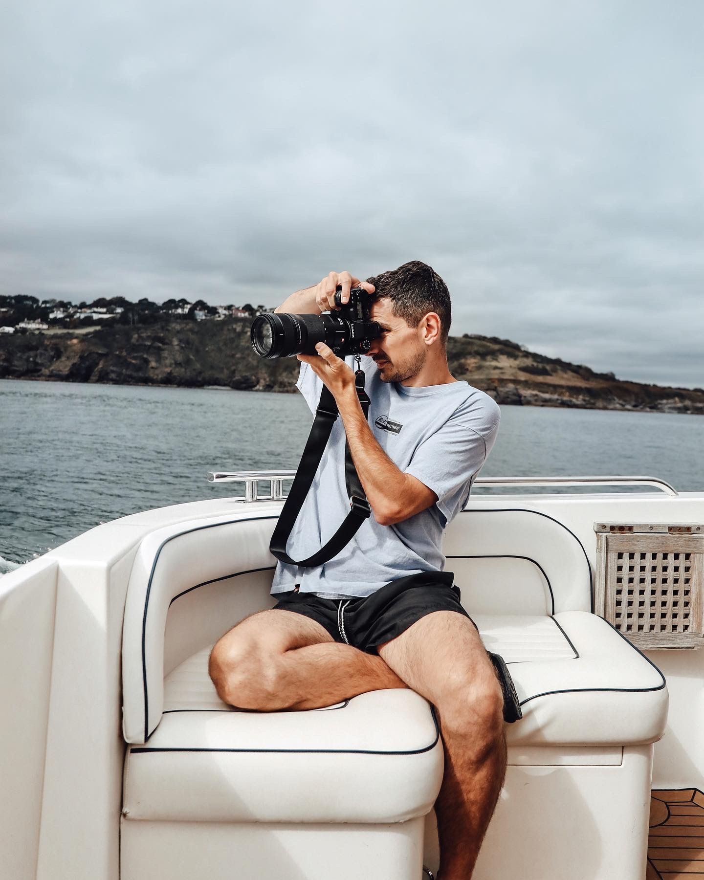 Photographer takes pictures from white boat cushioned seat with professional camera equipment against coastal backdrop.