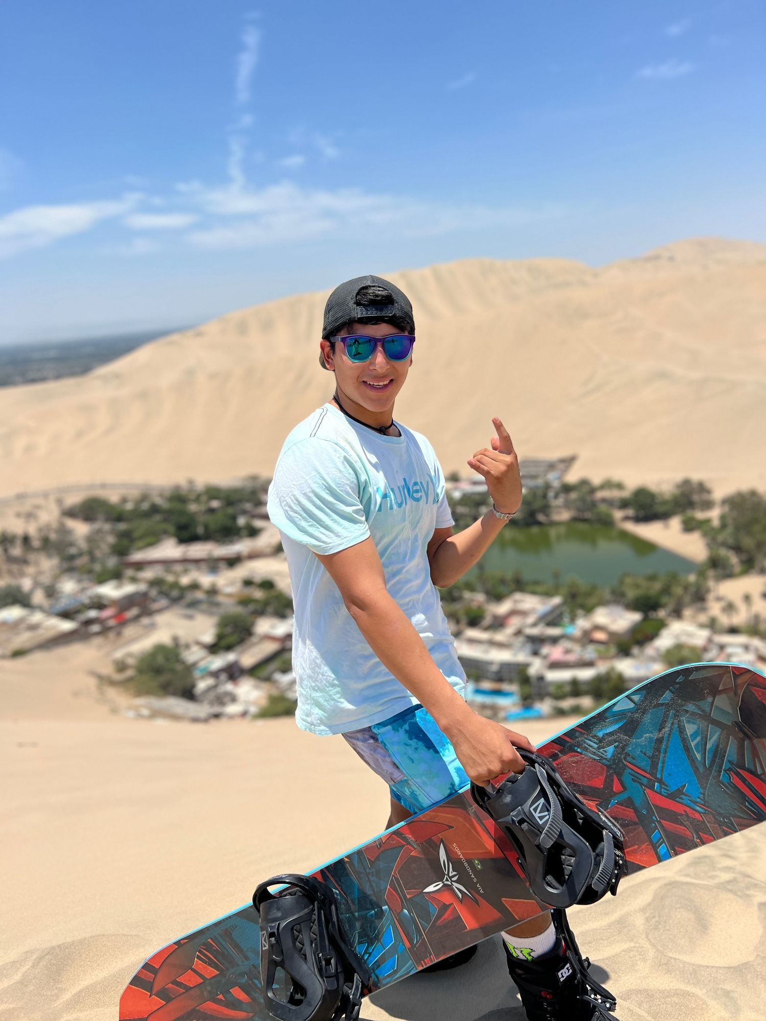 Sandboarder with sunglasses poses on dunes overlooking desert oasis landscape.