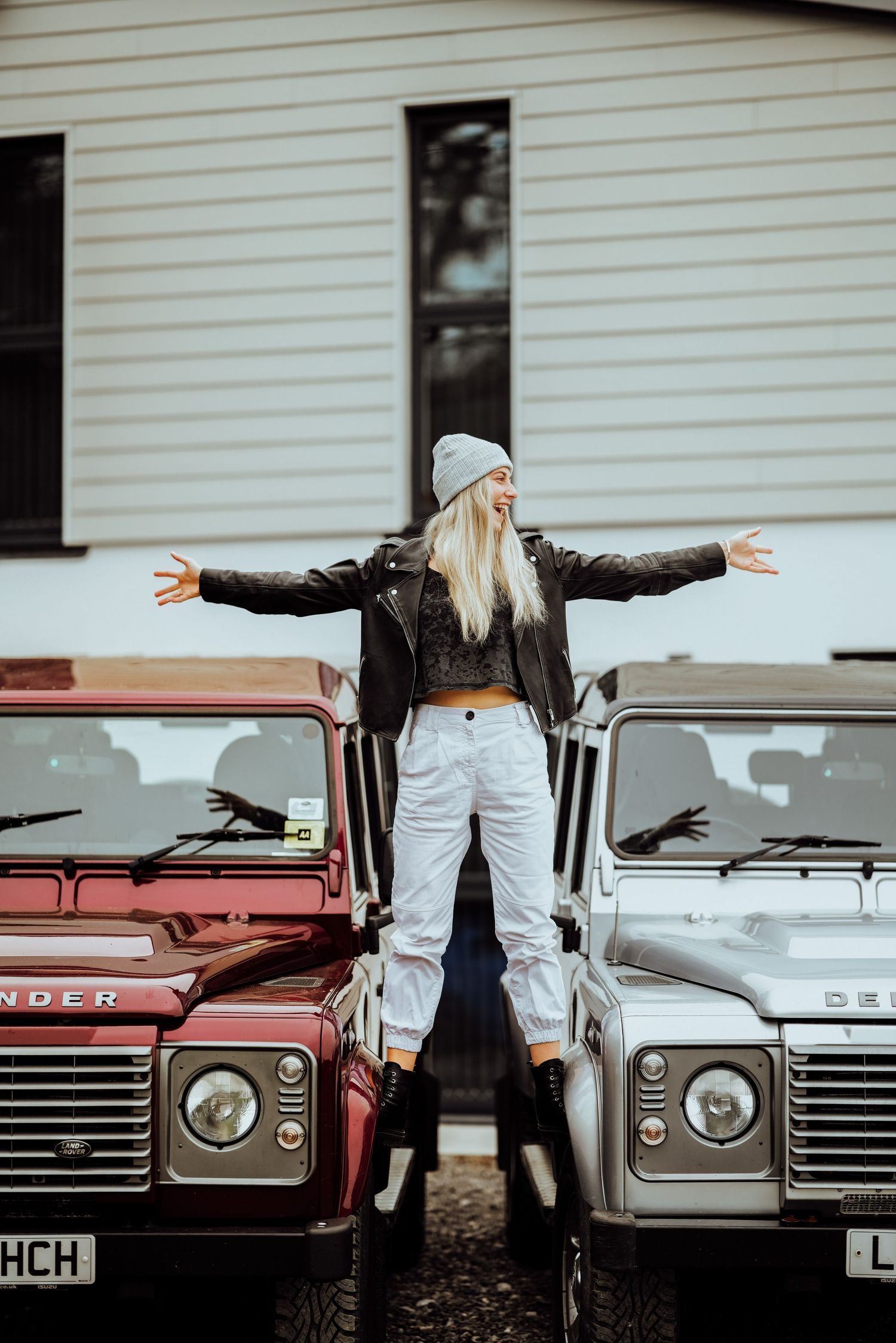 Person standing between two vintage Land Rover Defenders with arms outstretched wearing beanie and leather jacket.