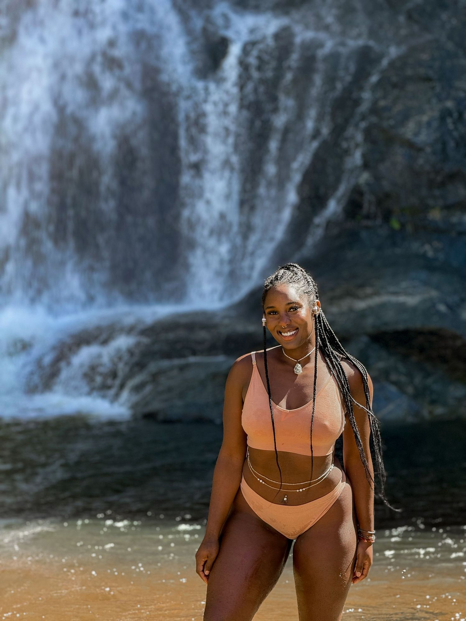 Woman in peach swimwear poses in front of a scenic waterfall on a sunny day.
