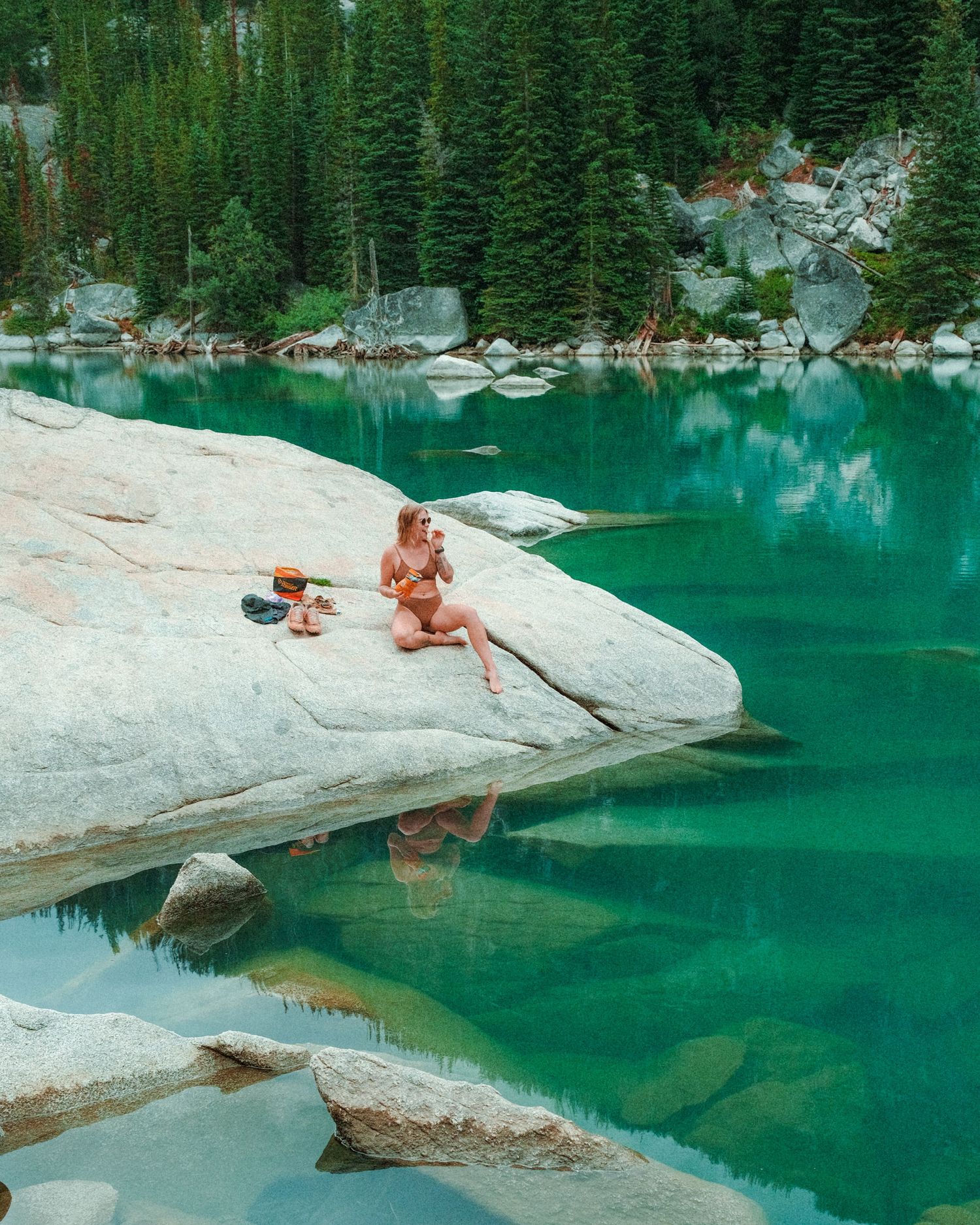 Someone relaxes on a white rock ledge beside a pristine turquoise alpine lake surrounded by evergreen forest.