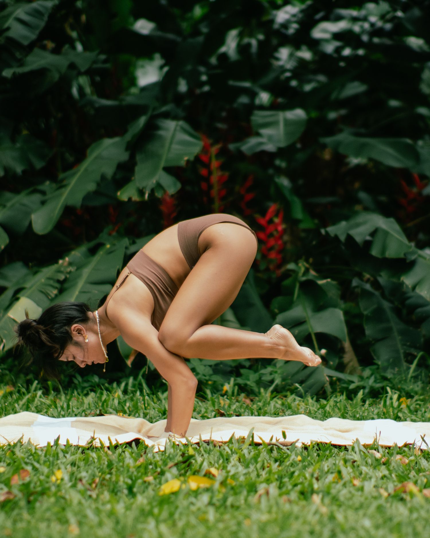 Yoga practitioner performs crow pose on a white mat outdoors surrounded by lush tropical foliage.