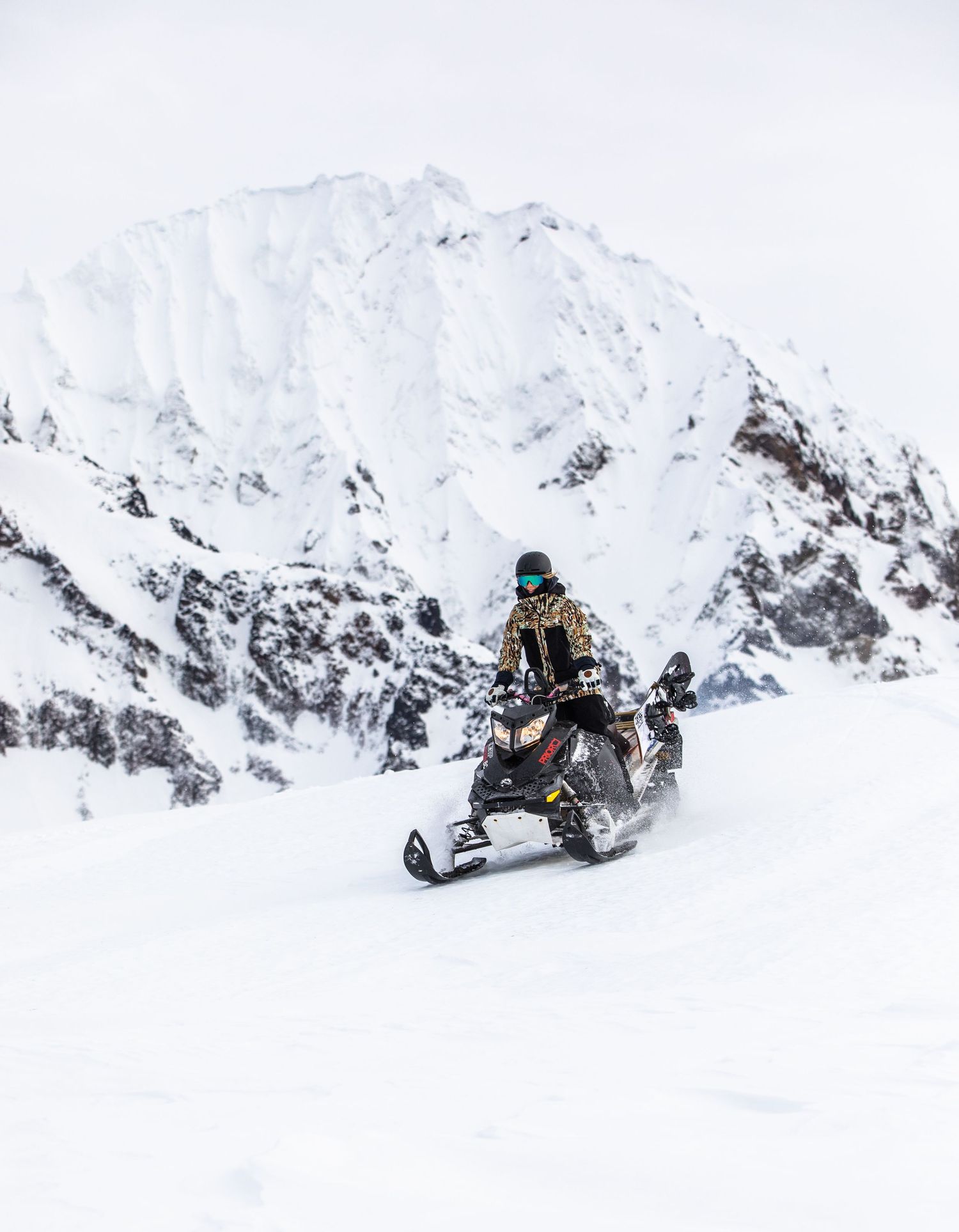 A snowmobiler rides across snowy terrain with towering white mountain peaks in the background.