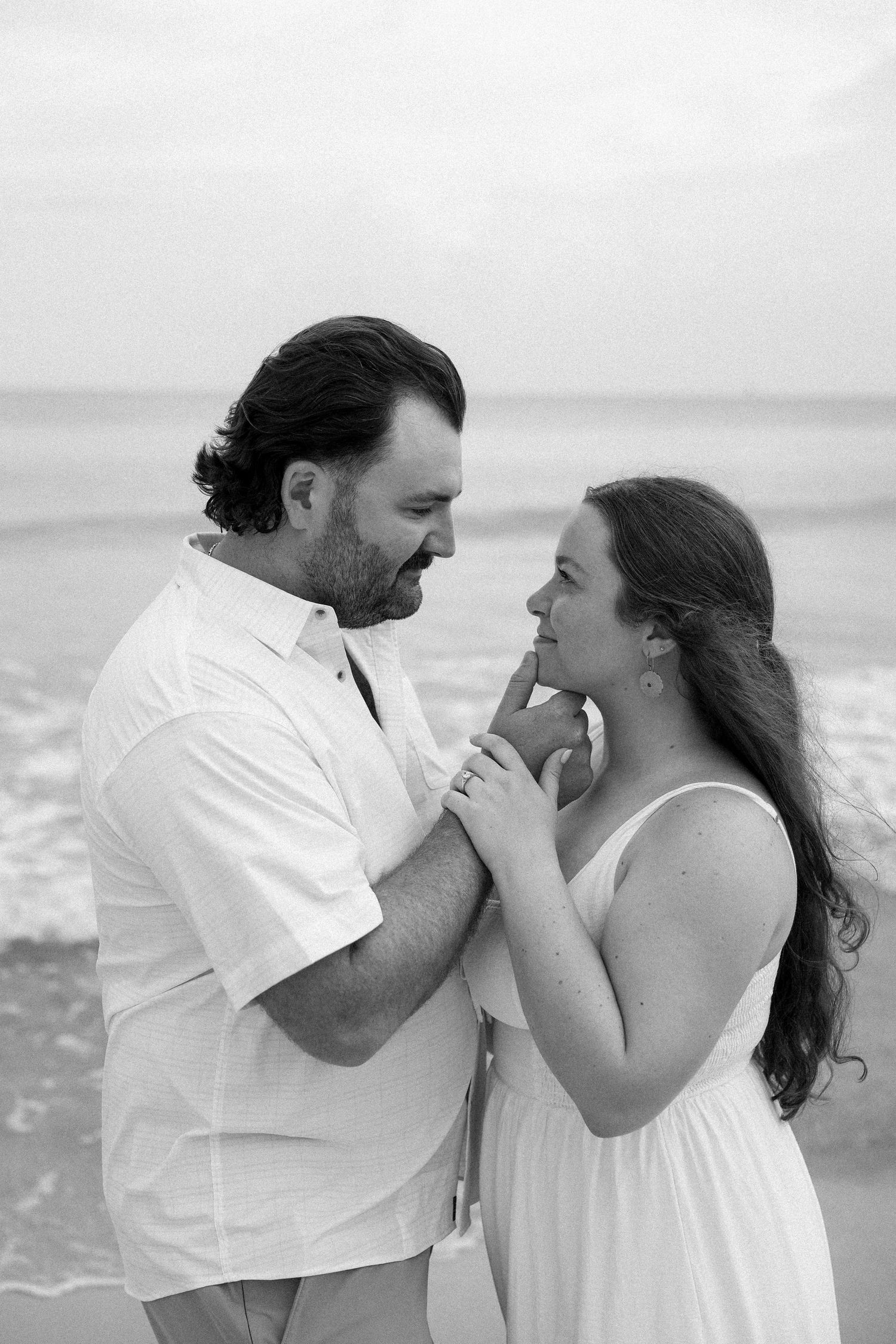 Black and white photo of a romantic couple sharing a tender moment on the beach, man gently touching womans chin.