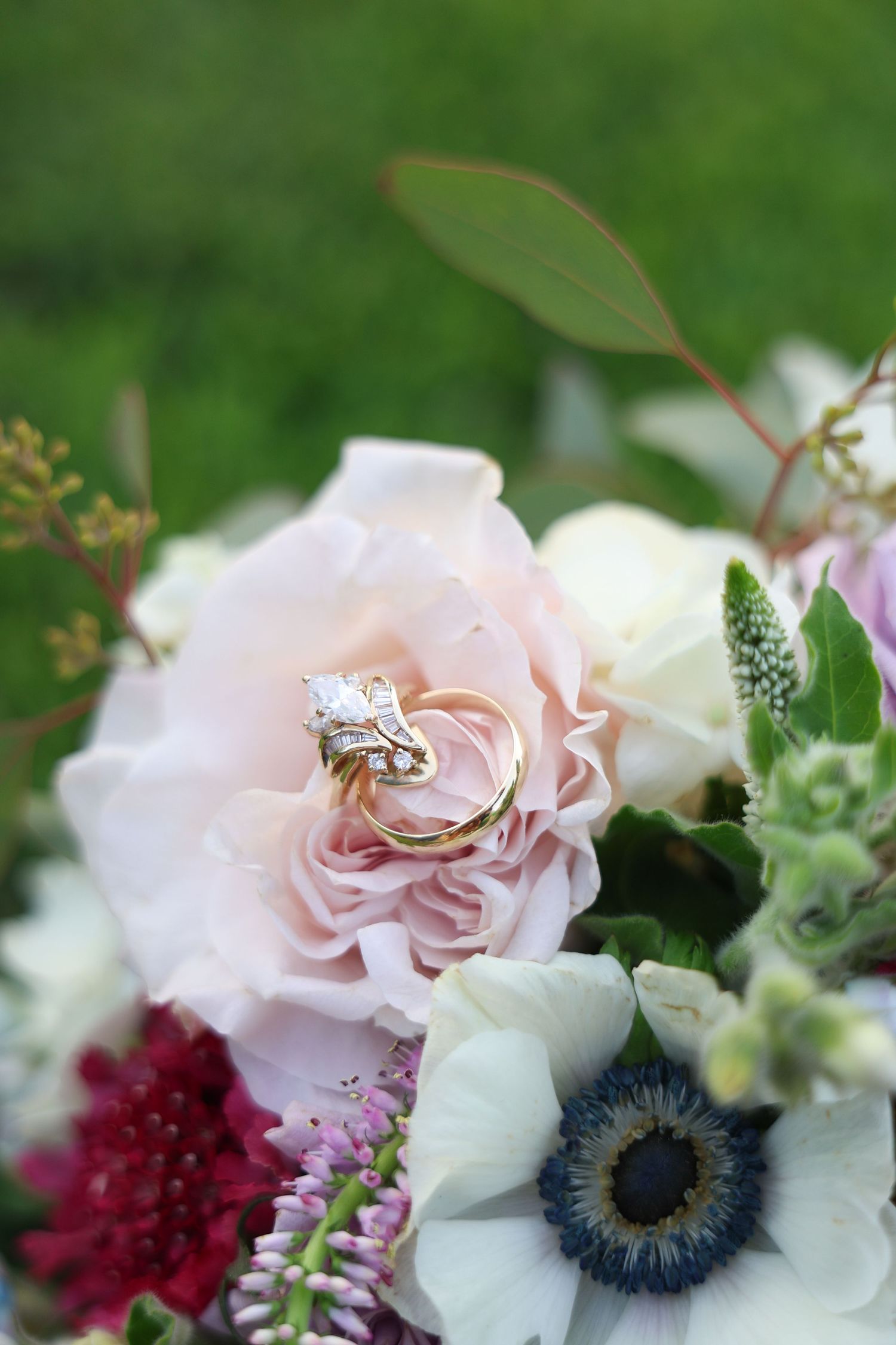 Gold wedding and engagement rings resting on a blush pink rose in a bridal bouquet with anemones and mixed florals.