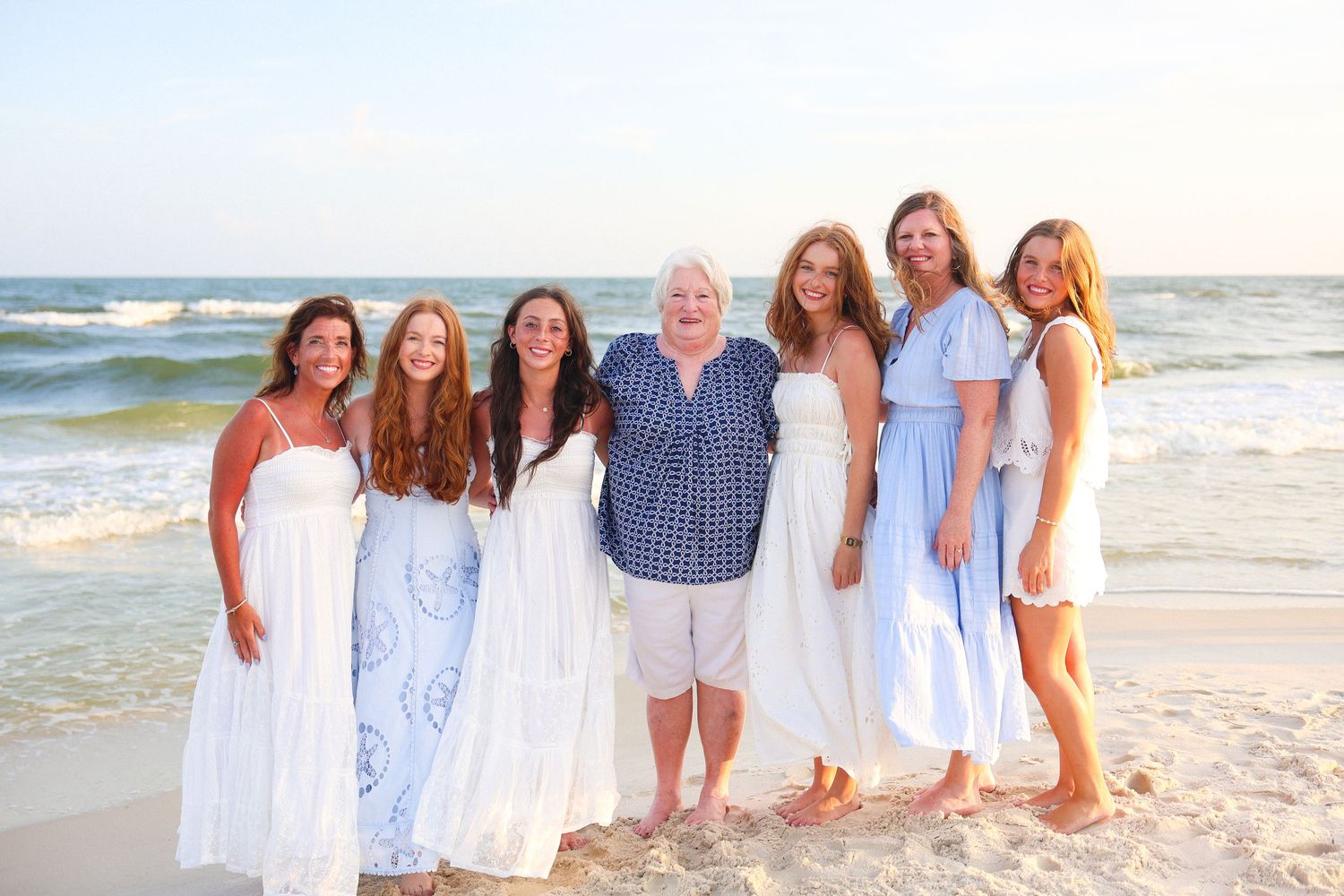 Seven women smiling together on a sunny beach, wearing white and blue outfits with ocean waves in the background.