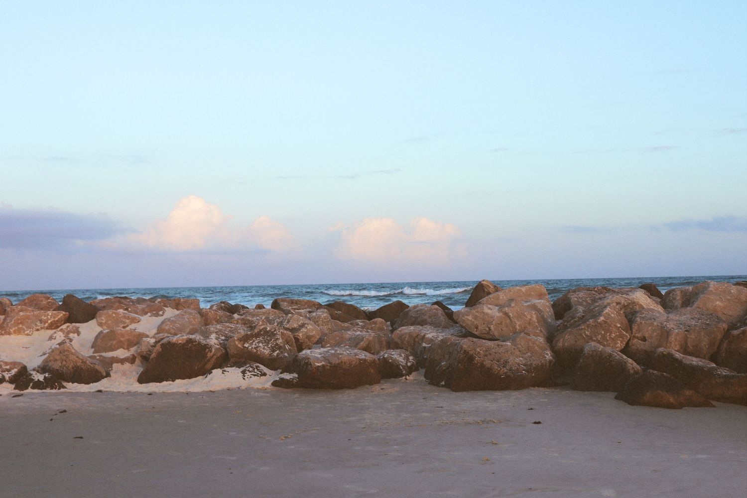 Rocky jetty on a tranquil sandy beach at dusk with soft clouds over the ocean horizon.