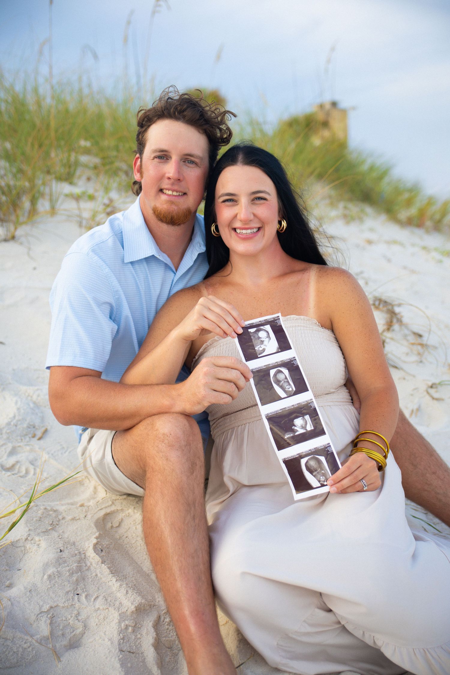 Happy couple sitting on beach sand dunes holding ultrasound photos, announcing pregnancy with joyful smiles.