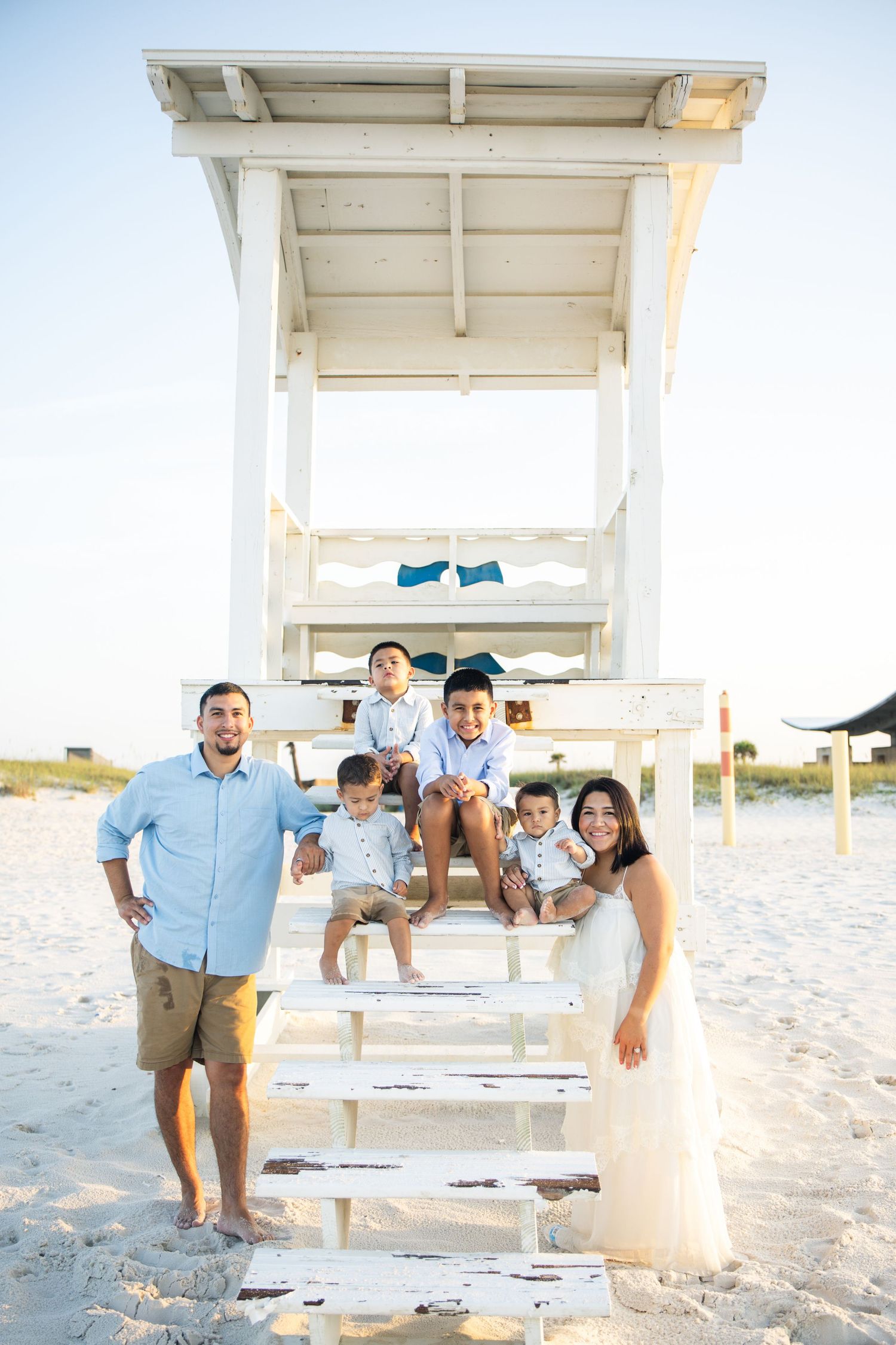 Family of five posing by a white lifeguard tower on a sunny beach, parents flanking three young boys on the steps.