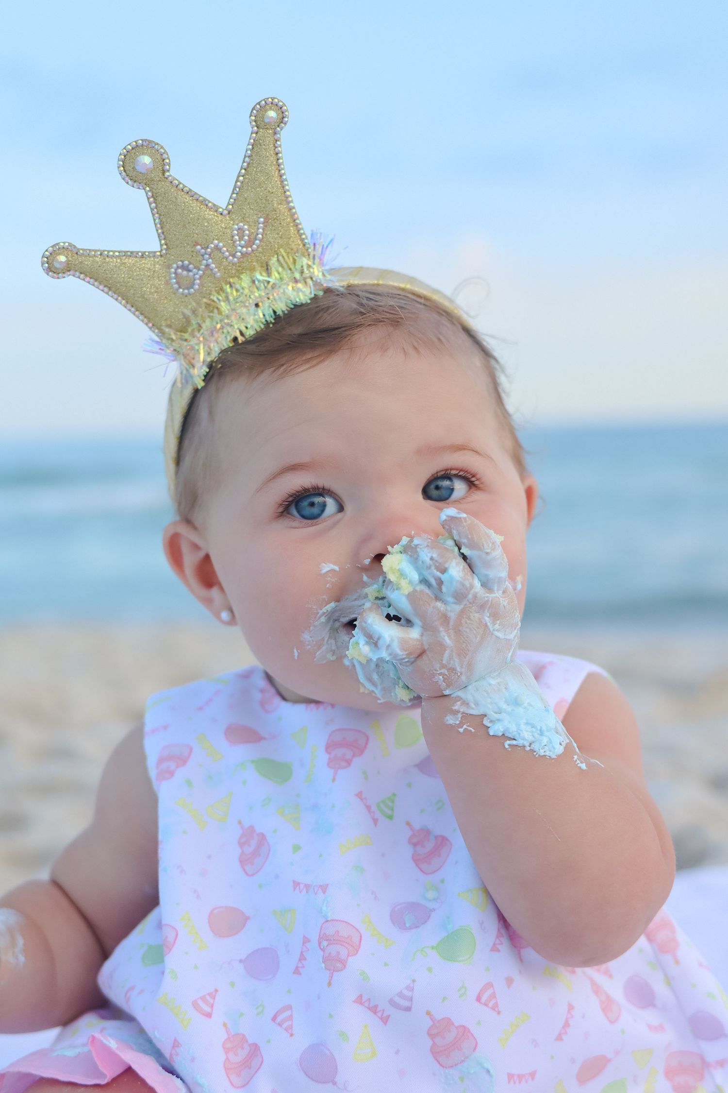 Adorable baby girl celebrating first birthday with cake smash on beach, wearing gold crown headband.
