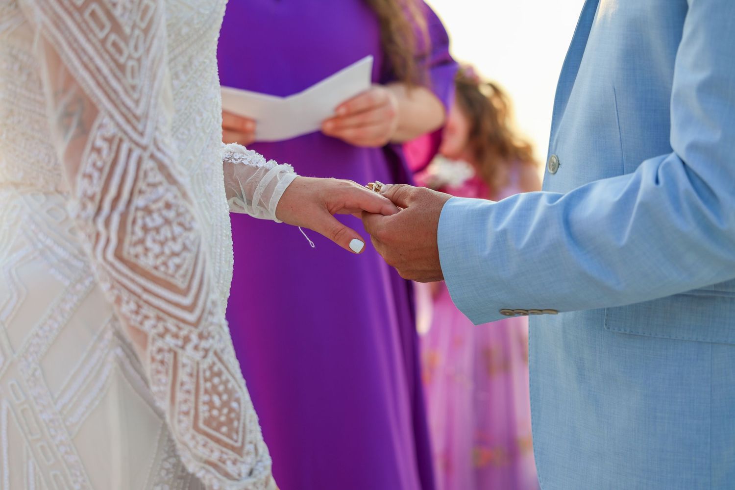 Groom in light blue suit placing wedding ring on bride's finger during outdoor ceremony with purple-dressed officiant.