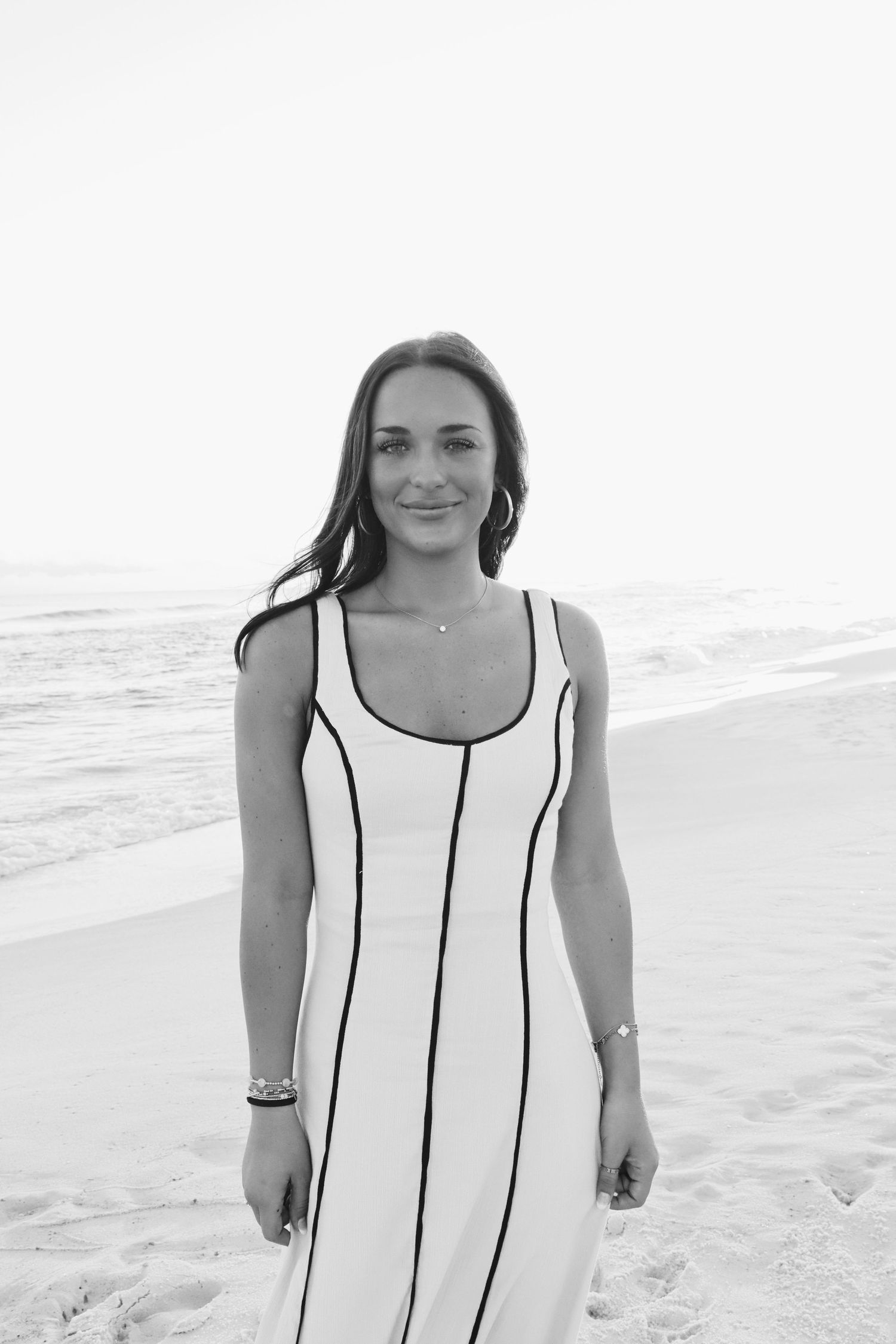 Brunette woman in white black-trimmed dress smiling softly on sandy beach in black and white.