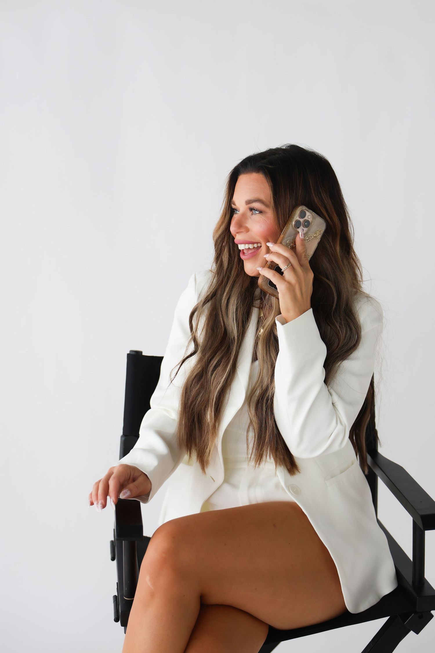 A smiling woman in a white blazer sits in a director's chair, talking on her smartphone against a white background.