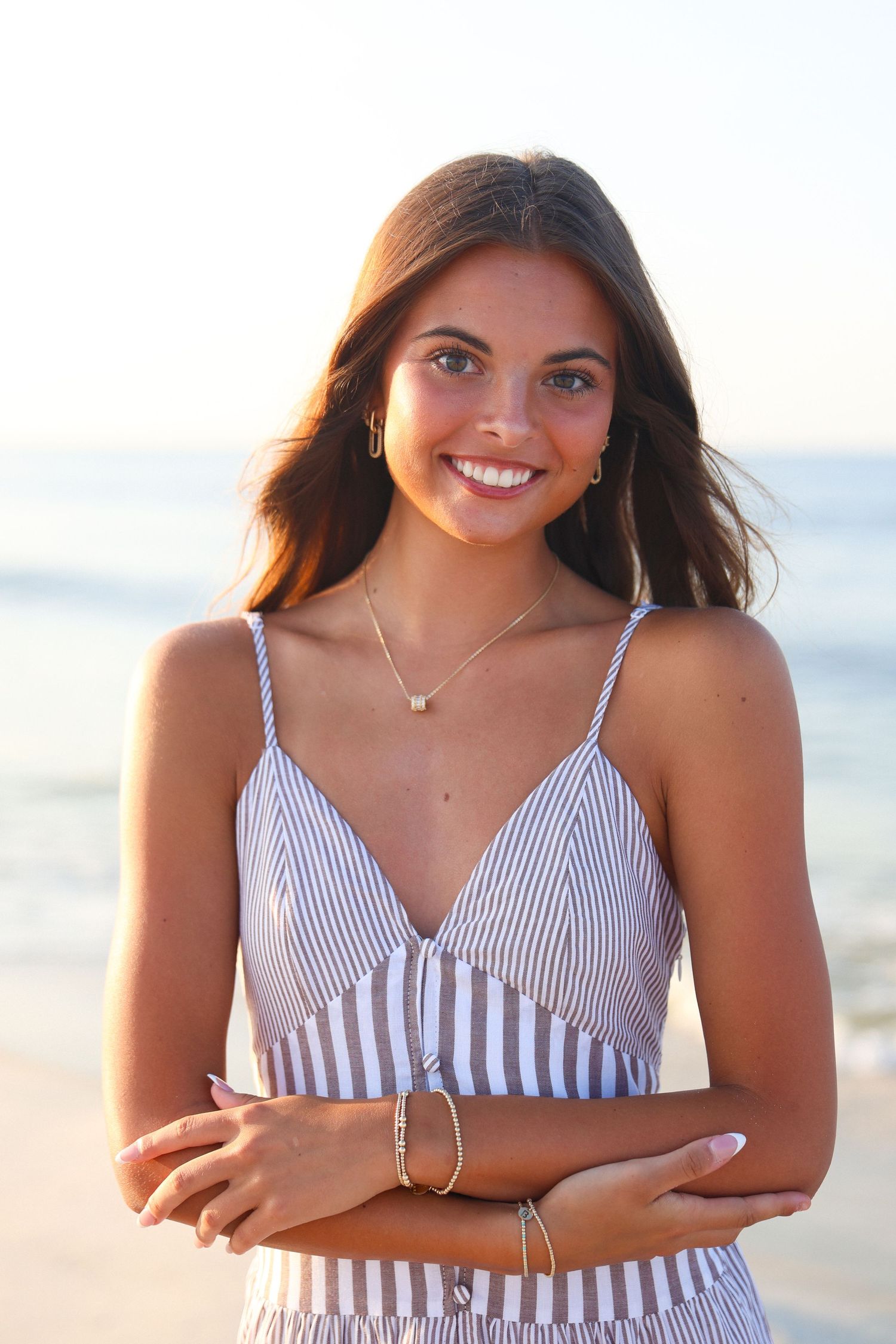 Smiling young woman in striped crop top with gold jewelry posing at beach during golden hour sunset.