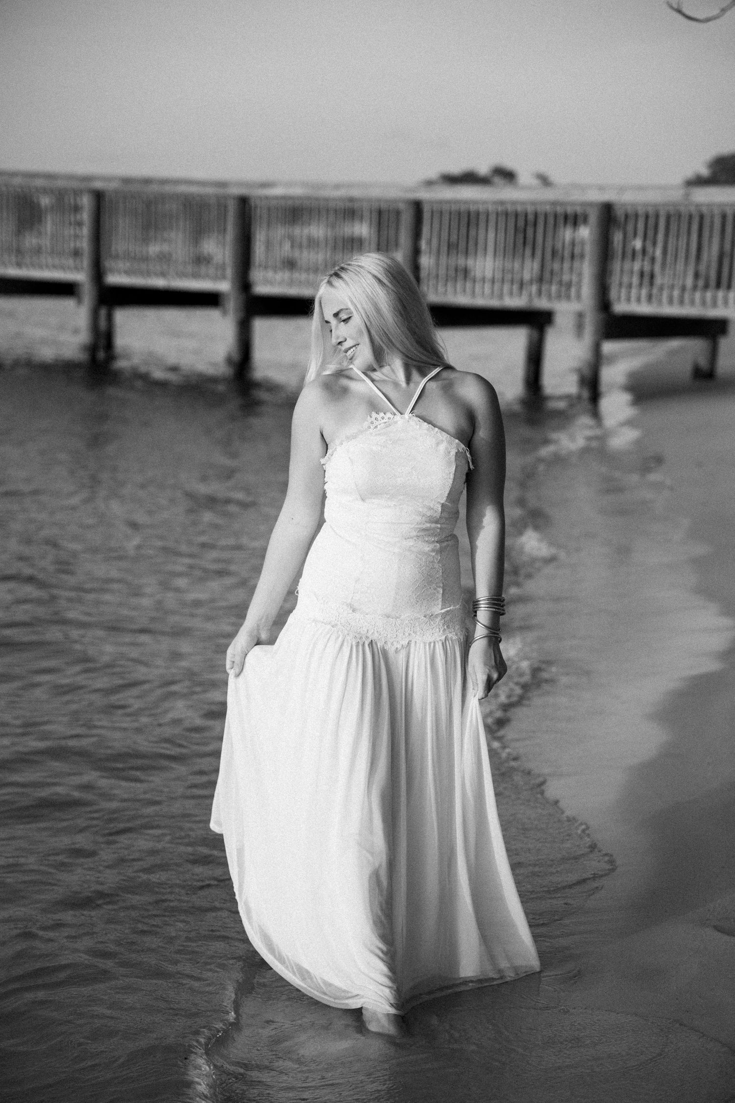 Blonde woman in white halter dress standing in shallow ocean water near a pier, black and white photo.