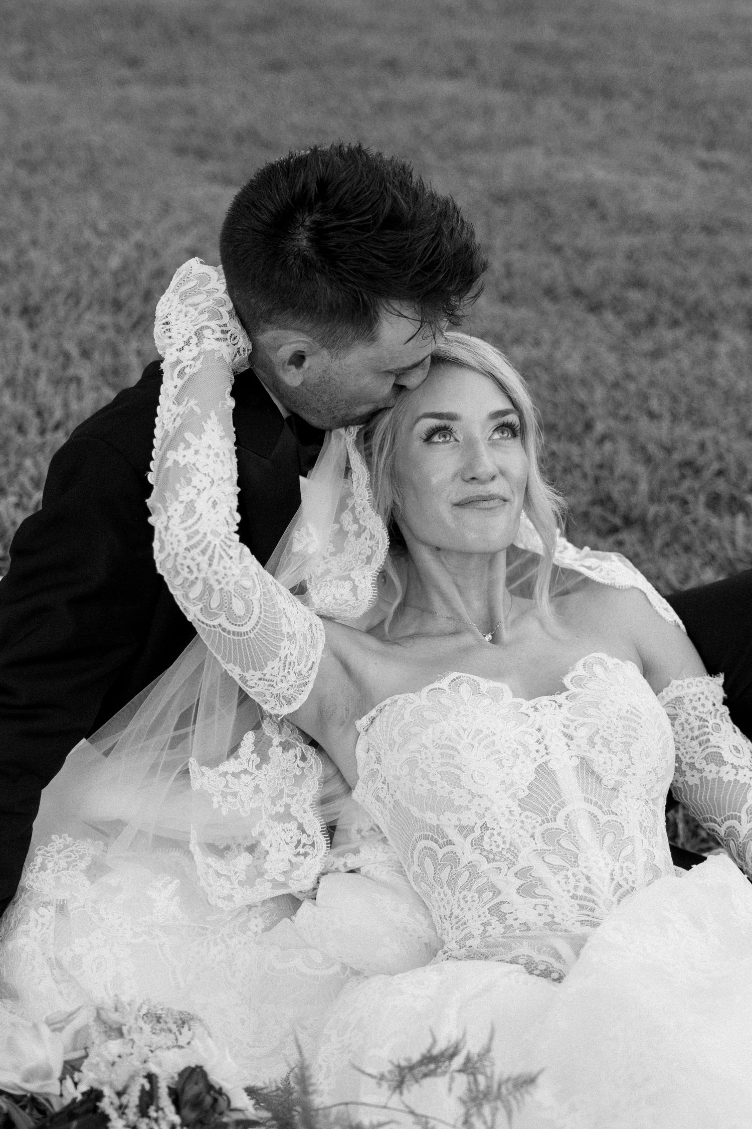 Black and white photo of a groom kissing bride on cheek while she smiles in lace wedding dress outdoors.