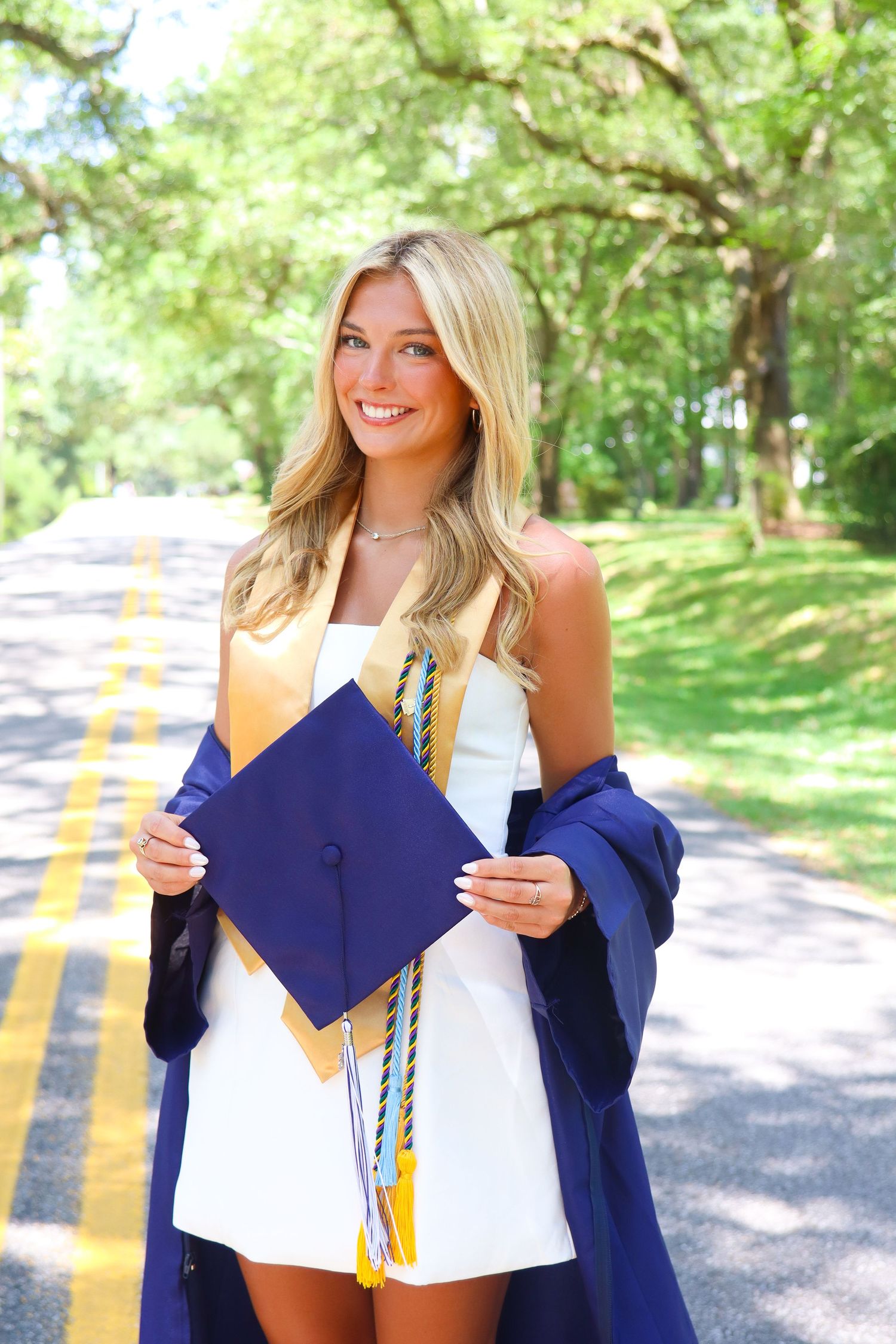 Smiling blonde graduate holding navy cap and gown with gold tassel on a tree-lined road in white dress.