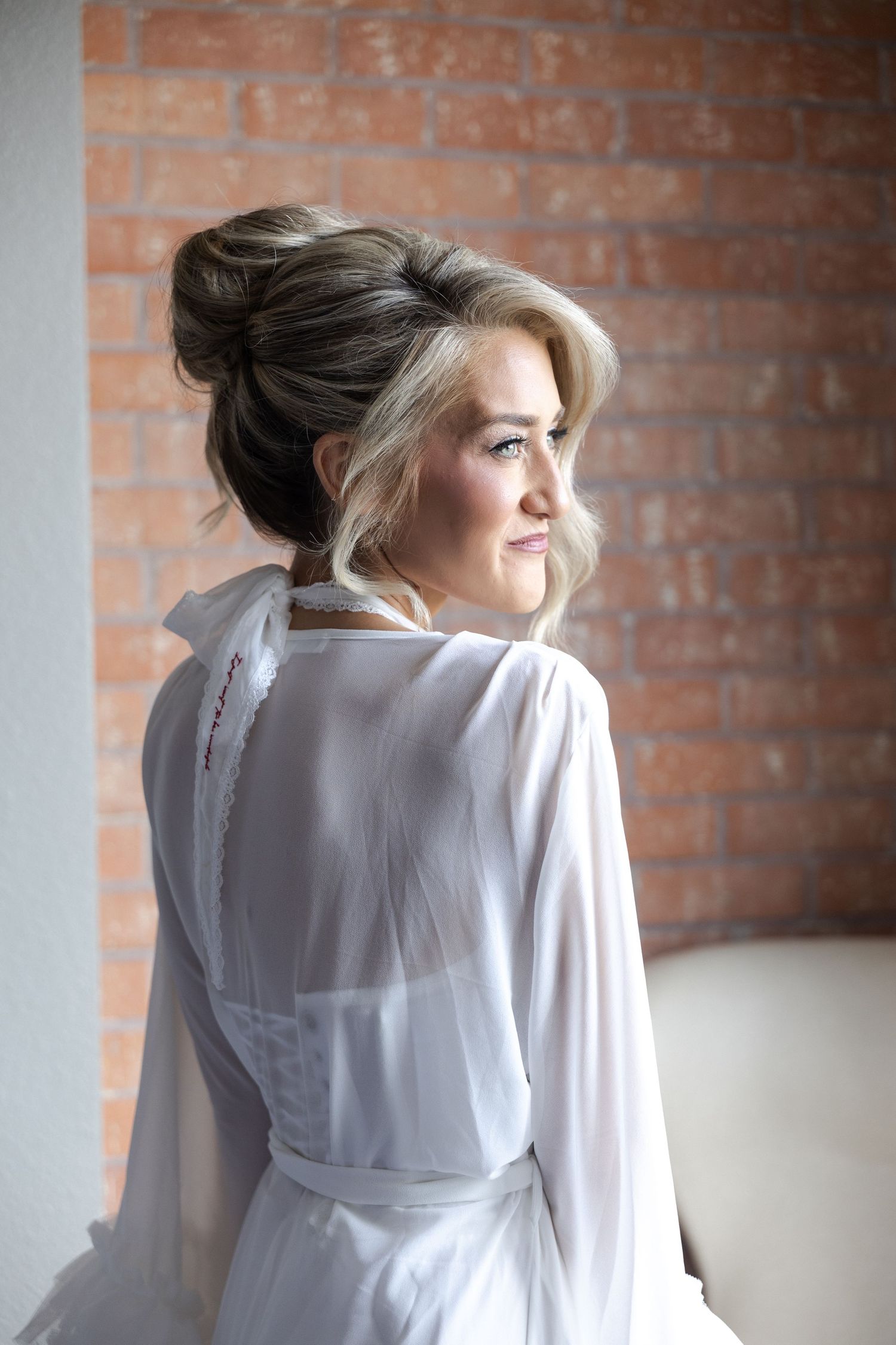 Elegant bride in white sheer robe with updo hairstyle, posing against a brick wall background.