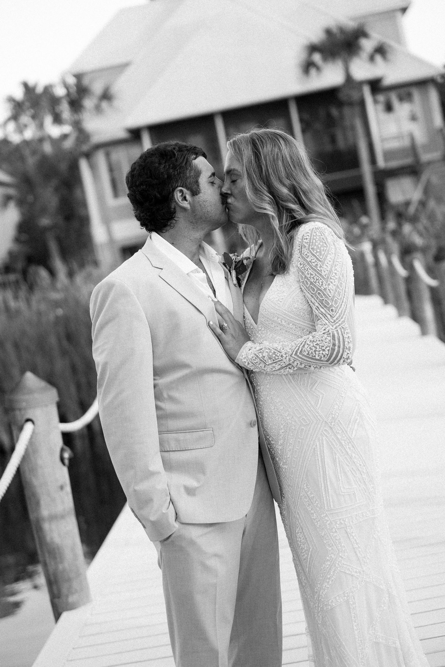 Black and white photo of a bride and groom sharing a kiss on a dock, with a waterfront building in the background.