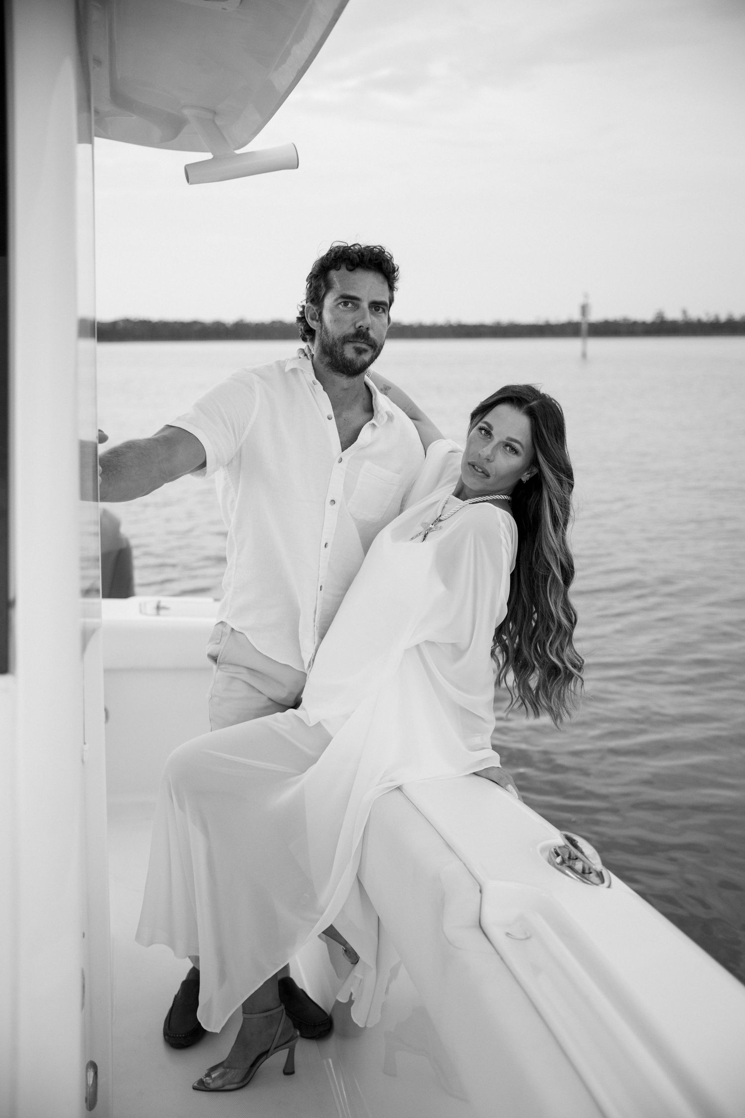 Black and white photo of an elegant couple in white outfits posing on a luxury boat with water in background.