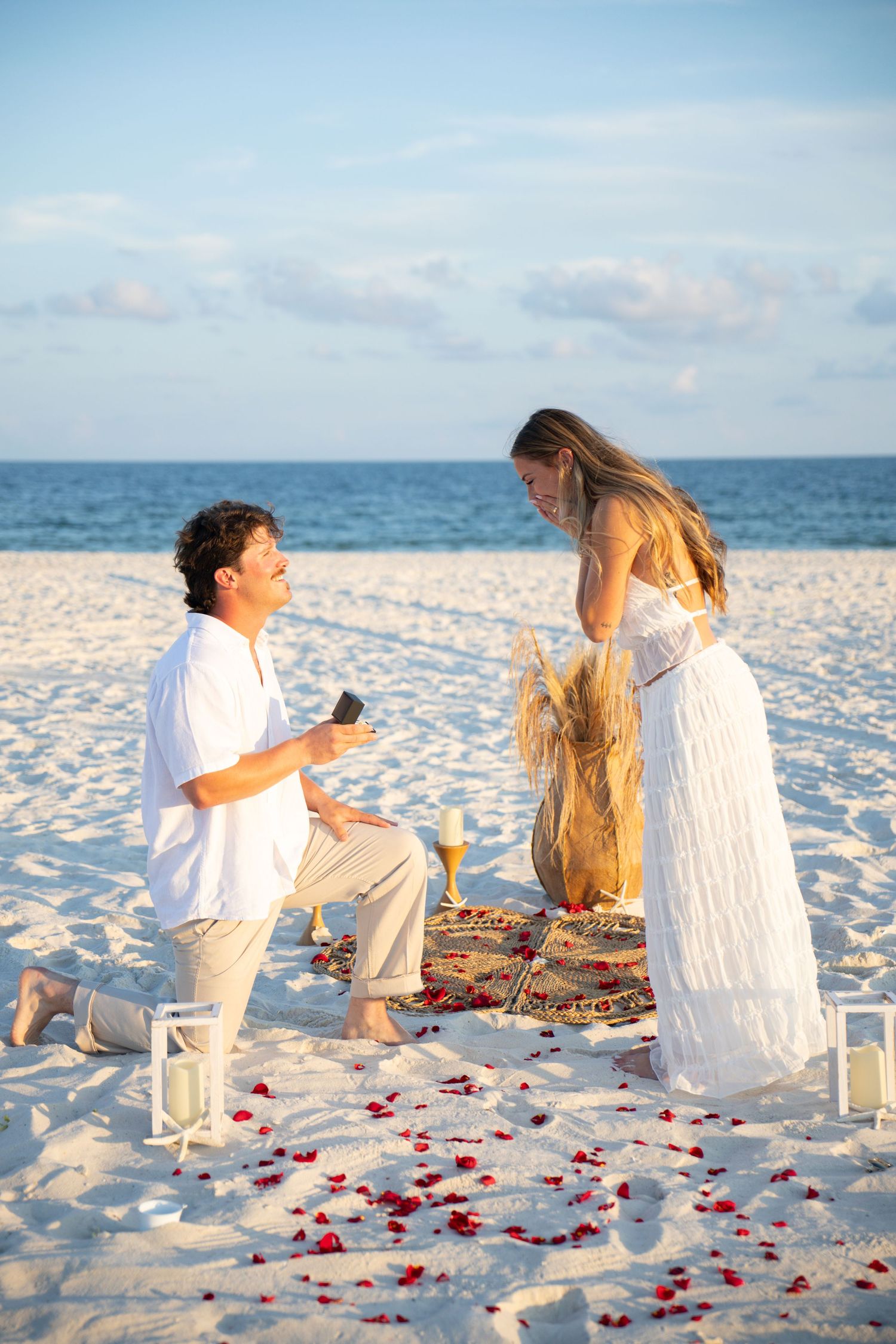 Man proposes to woman on one knee on a romantic beach with rose petals, lanterns, and champagne at sunset.