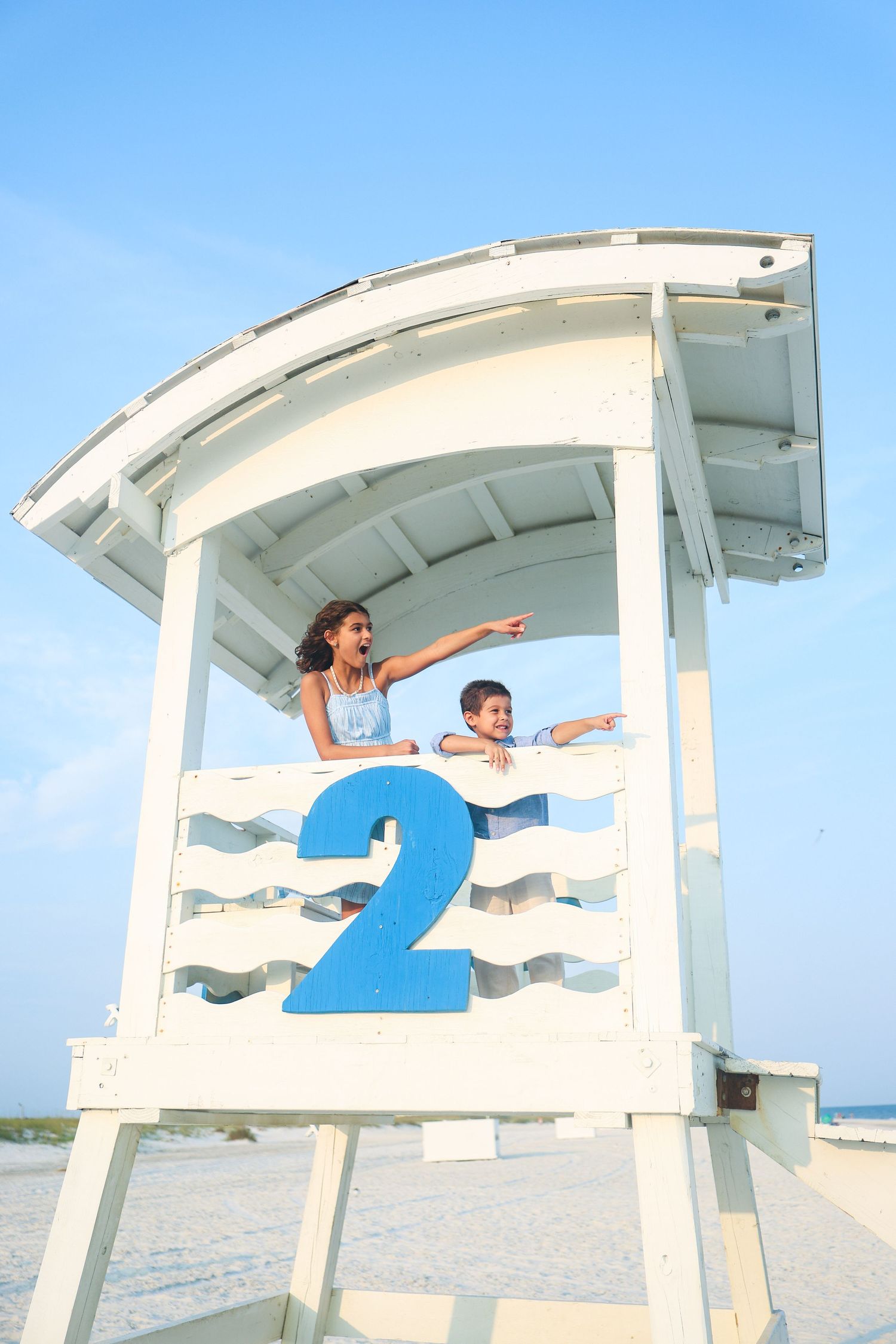 Two children point excitedly from a white beach lifeguard station number 2 on a sunny day.
