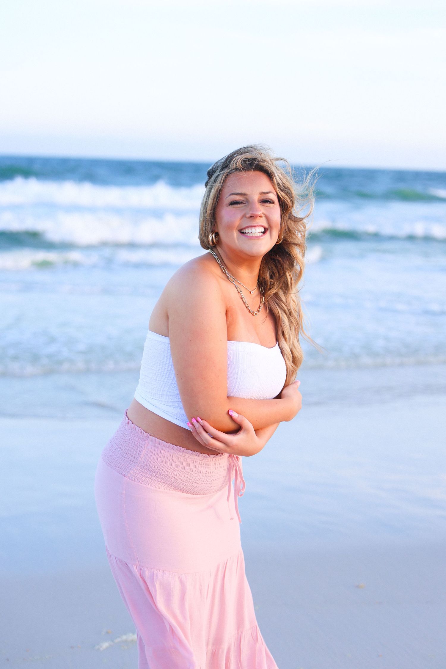 Smiling blonde woman in white crop top and pink maxi skirt posing on a sandy beach with ocean waves behind her.