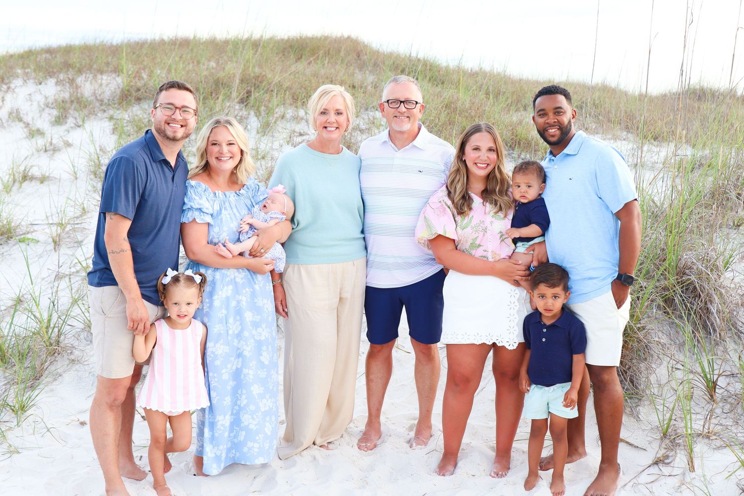 Multi-generational family posing together on a sandy beach with dunes, dressed in coordinated blue and white outfits.