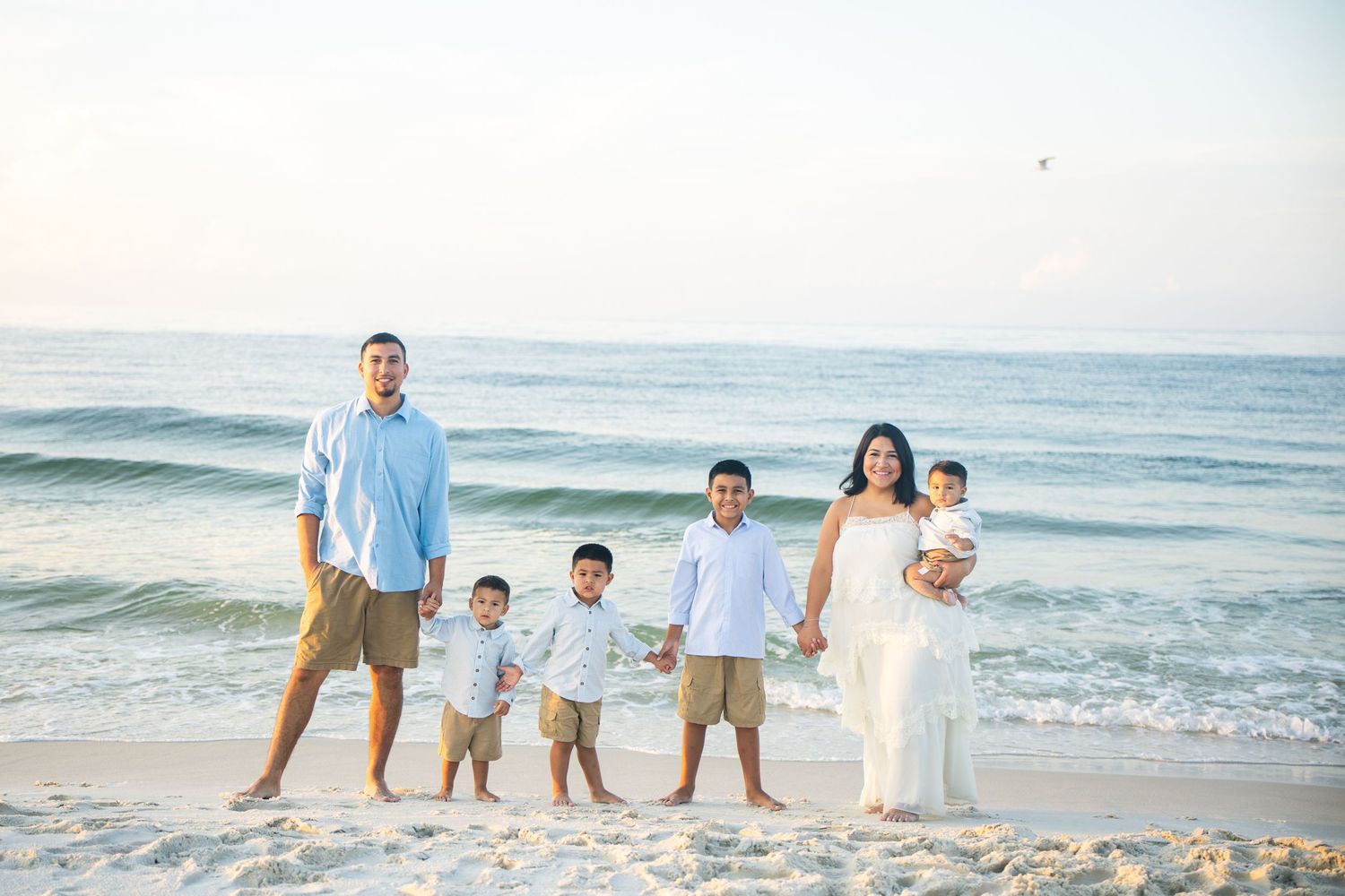 Family of six poses on a sandy beach with ocean waves, wearing coordinated light blue and white outfits.