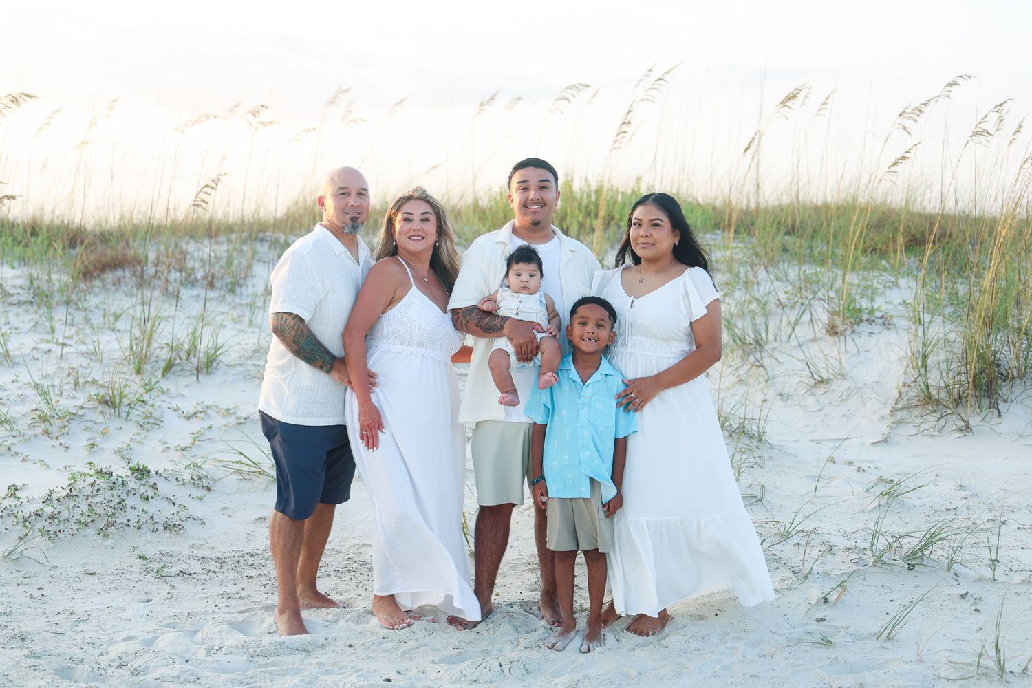 Family of six posing on sandy beach with sea oats, wearing white and light blue outfits at golden hour.