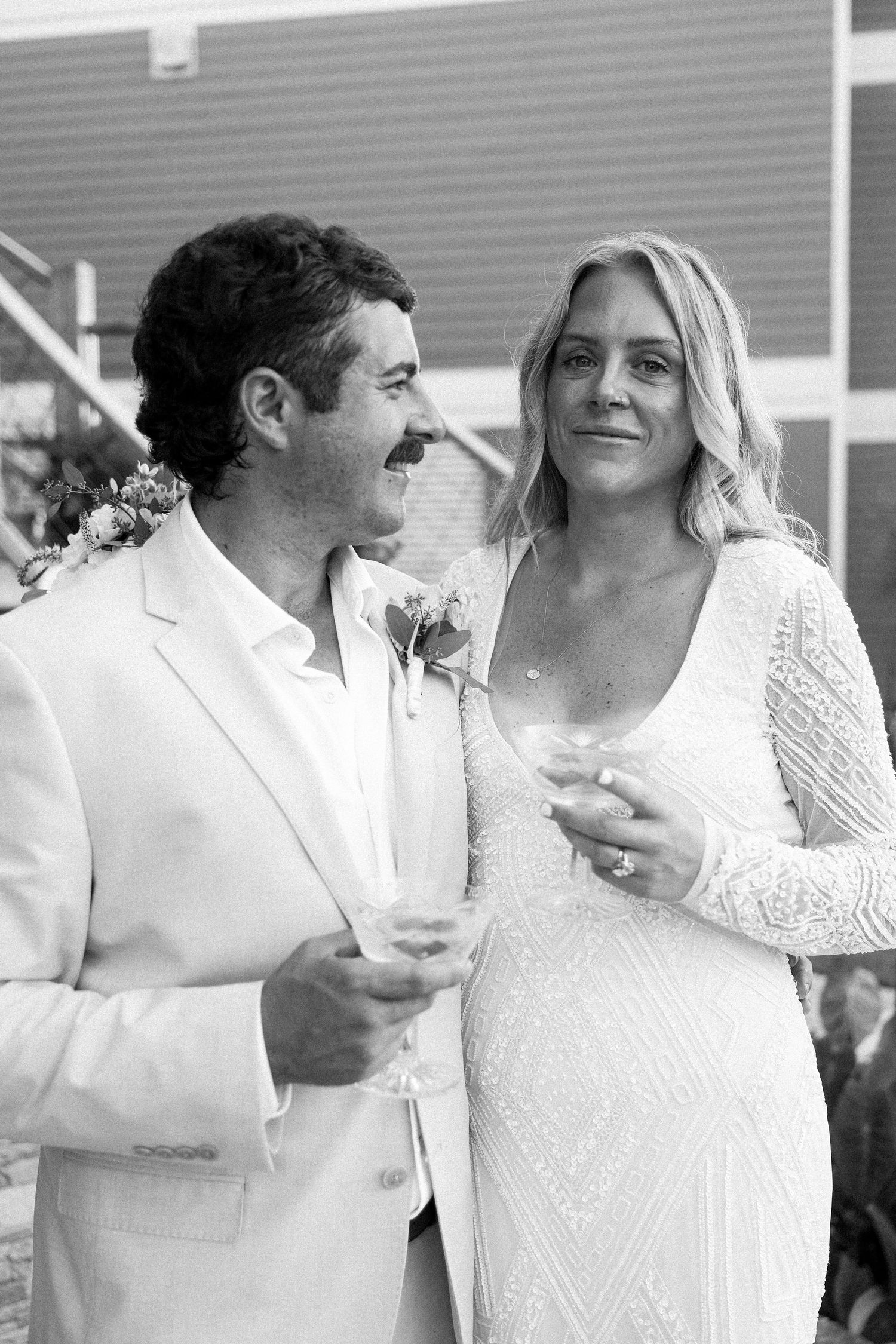 Black and white photo of a smiling bride in lace gown and groom in white suit holding champagne glasses at wedding.