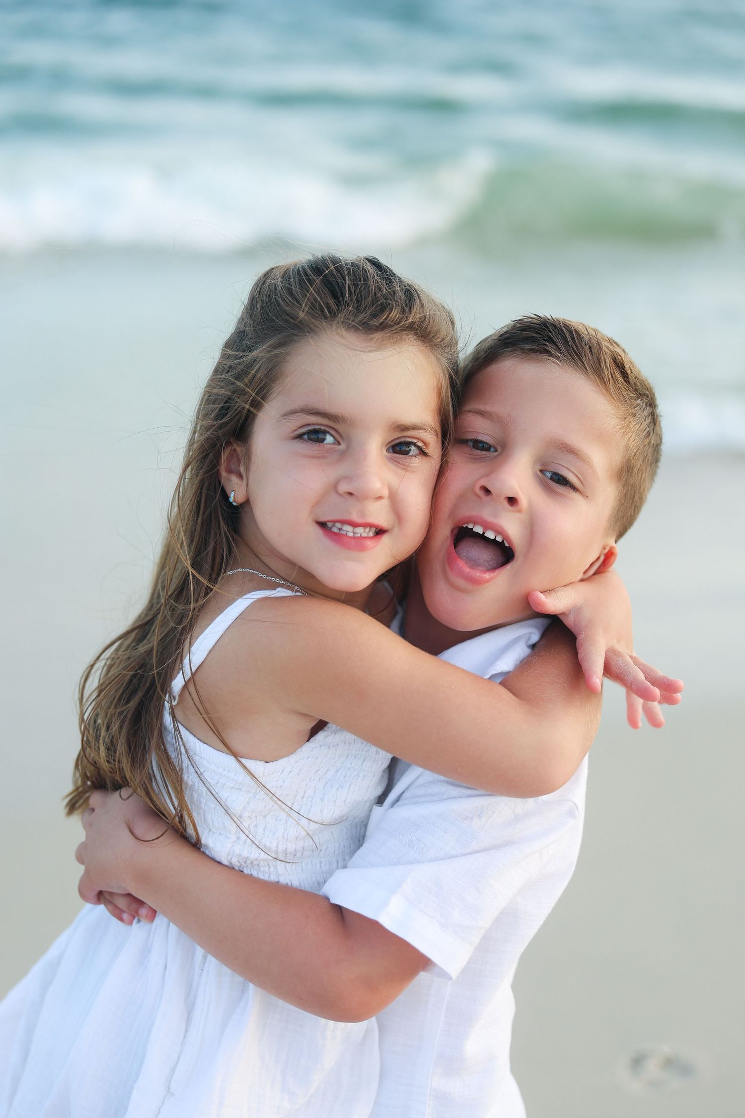 Two happy children hugging and smiling on a sandy beach with ocean waves in the background.