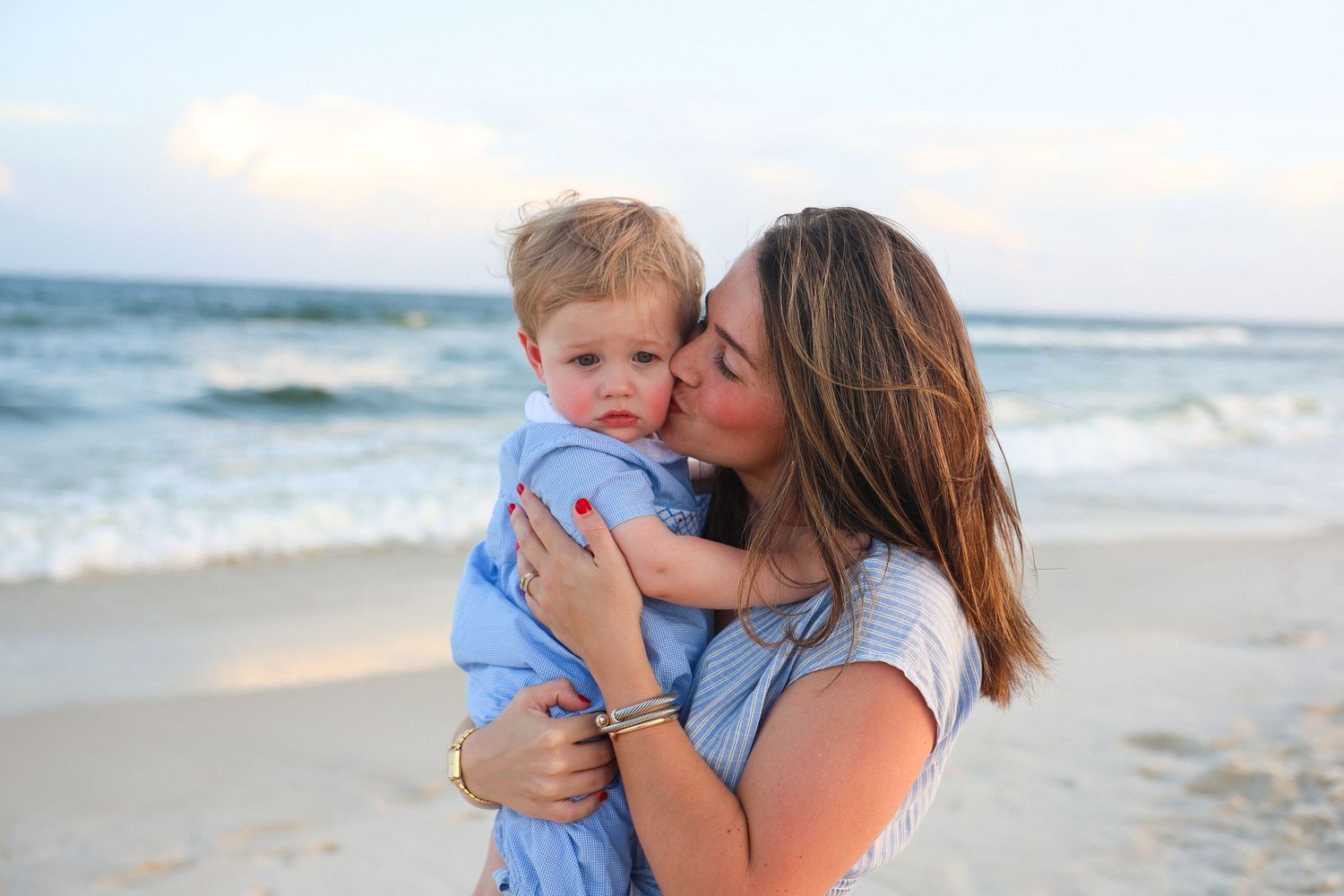 A mother kissing her toddler son on the cheek while holding him on a sandy beach near the ocean.