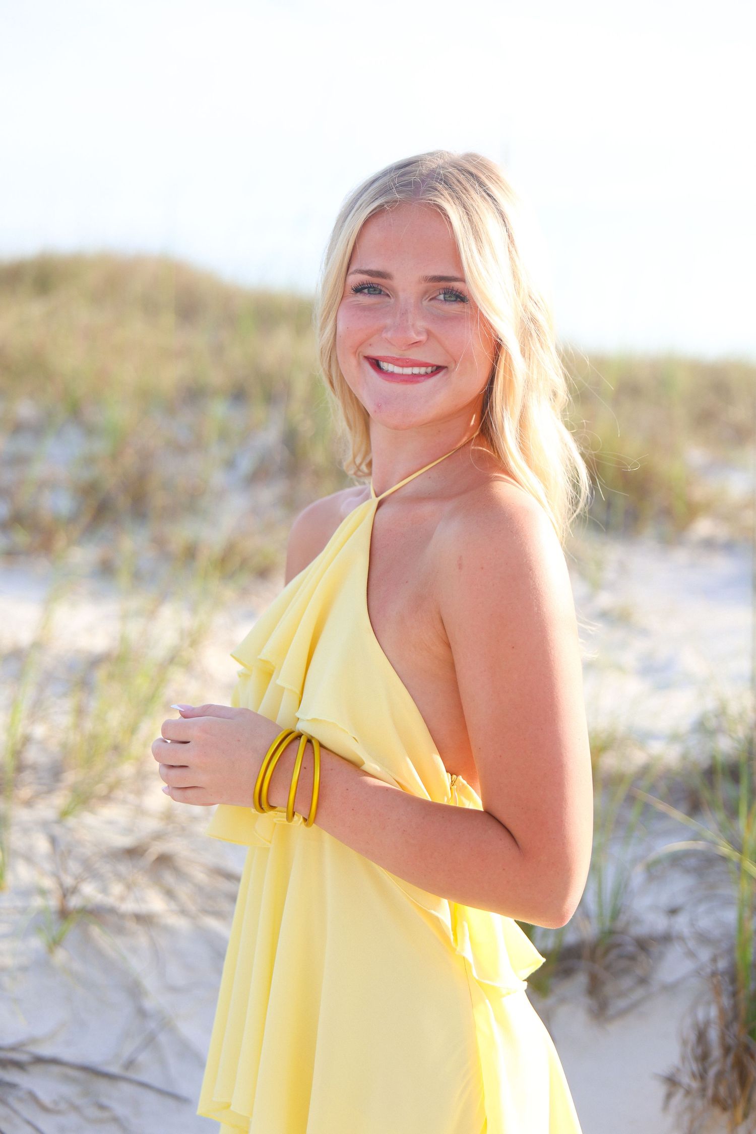 Smiling blonde woman in yellow sundress posing on sandy beach dunes in bright sunlight.