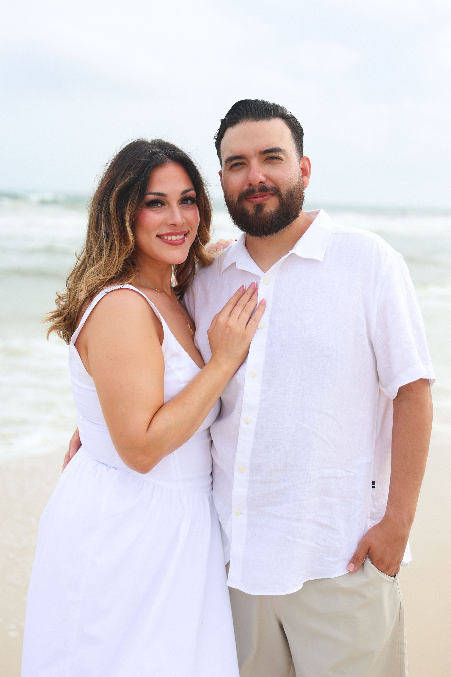 Couple in white outfits posing together on a beach with ocean waves in the background.