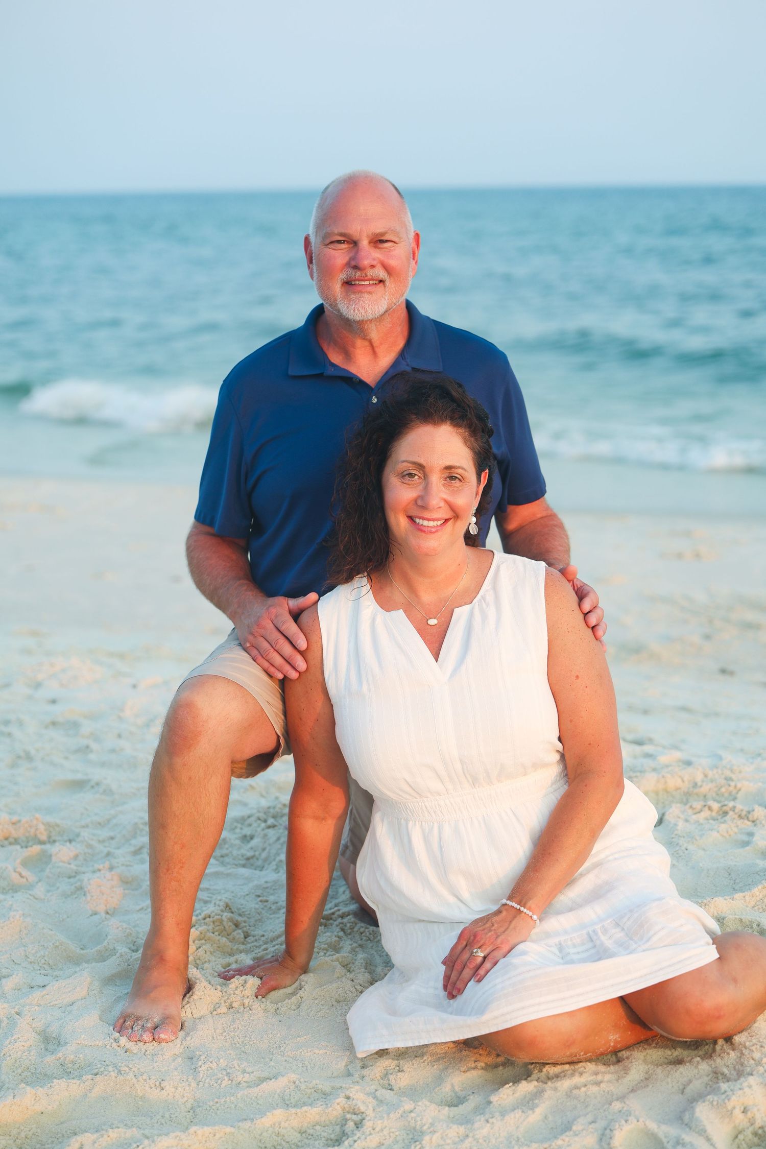 Smiling middle-aged couple posing on sandy beach with ocean waves, man in blue polo, woman in white dress.