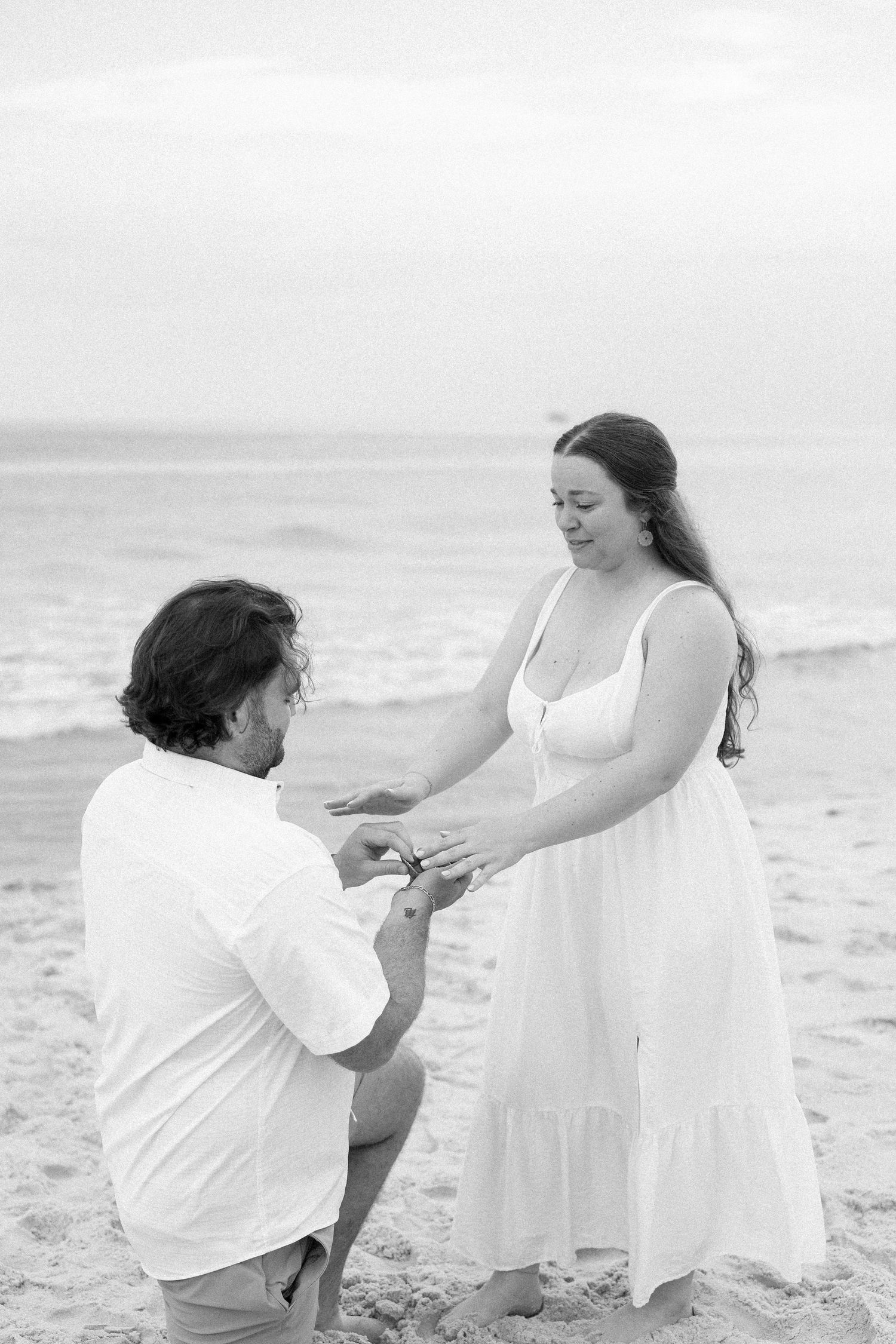 Black and white beach proposal photo of man on one knee placing ring on womans finger, ocean waves in background.