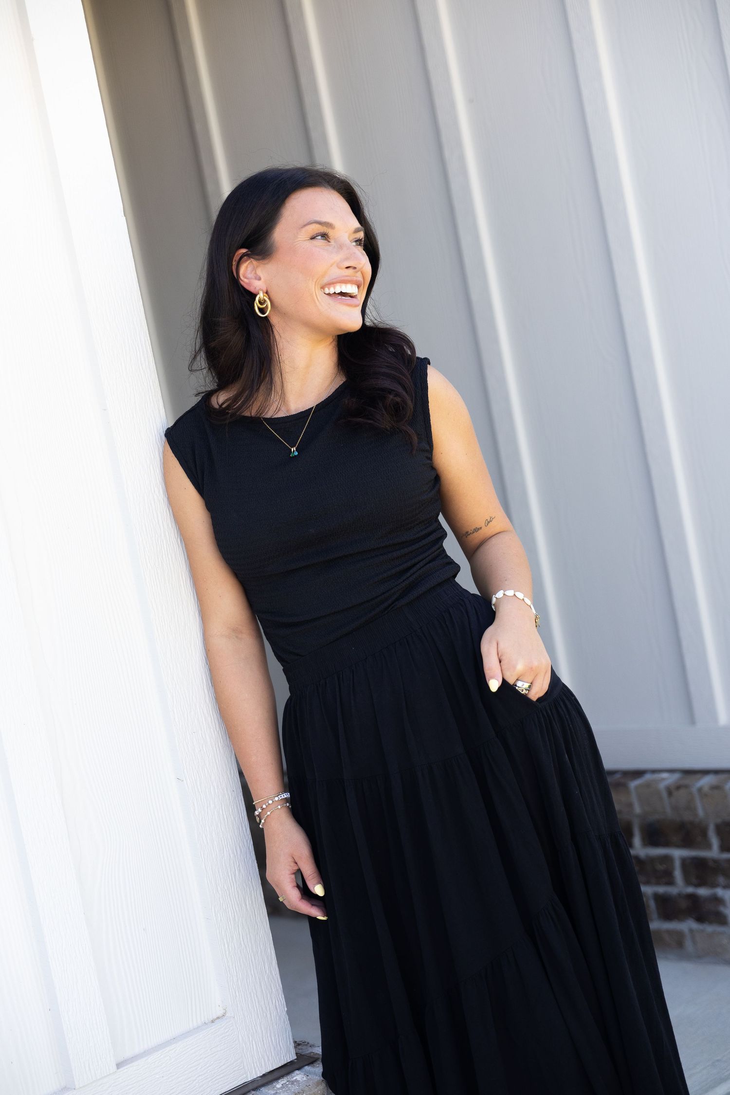 Woman in black sleeveless maxi dress laughing outdoors, wearing gold earrings and delicate necklace.