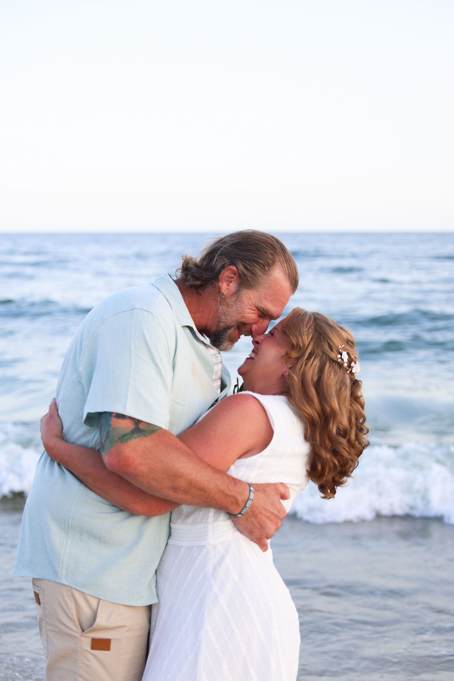 Couple sharing a romantic moment on the beach, dressed in white and light blue, with ocean waves in the background.